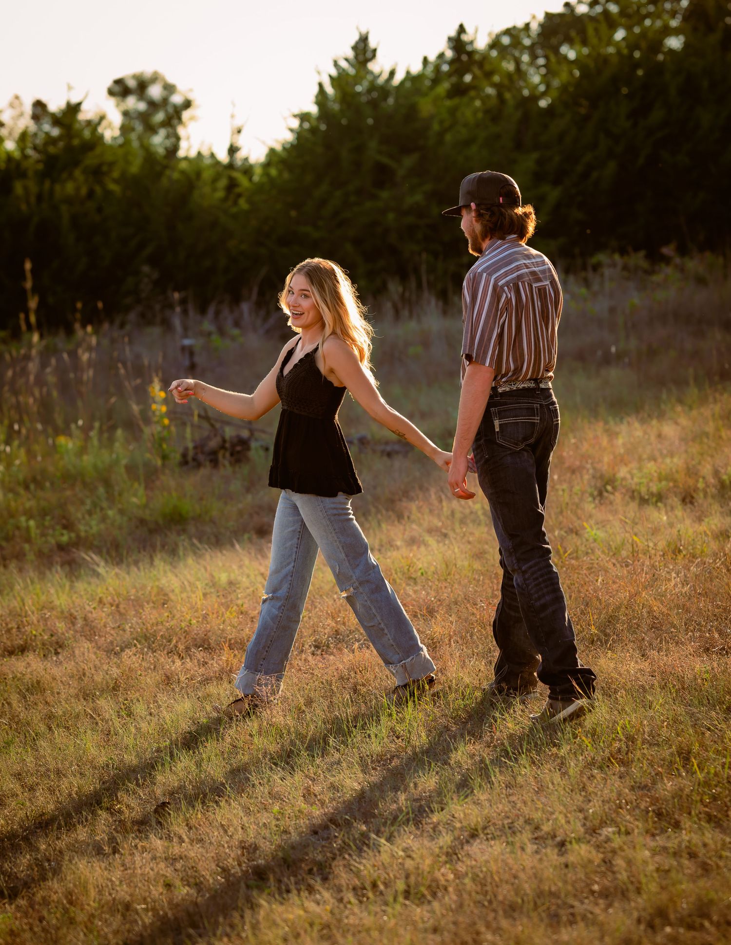 Young couple holding hands and strolling through a sun-drenched grassy field at golden hour.