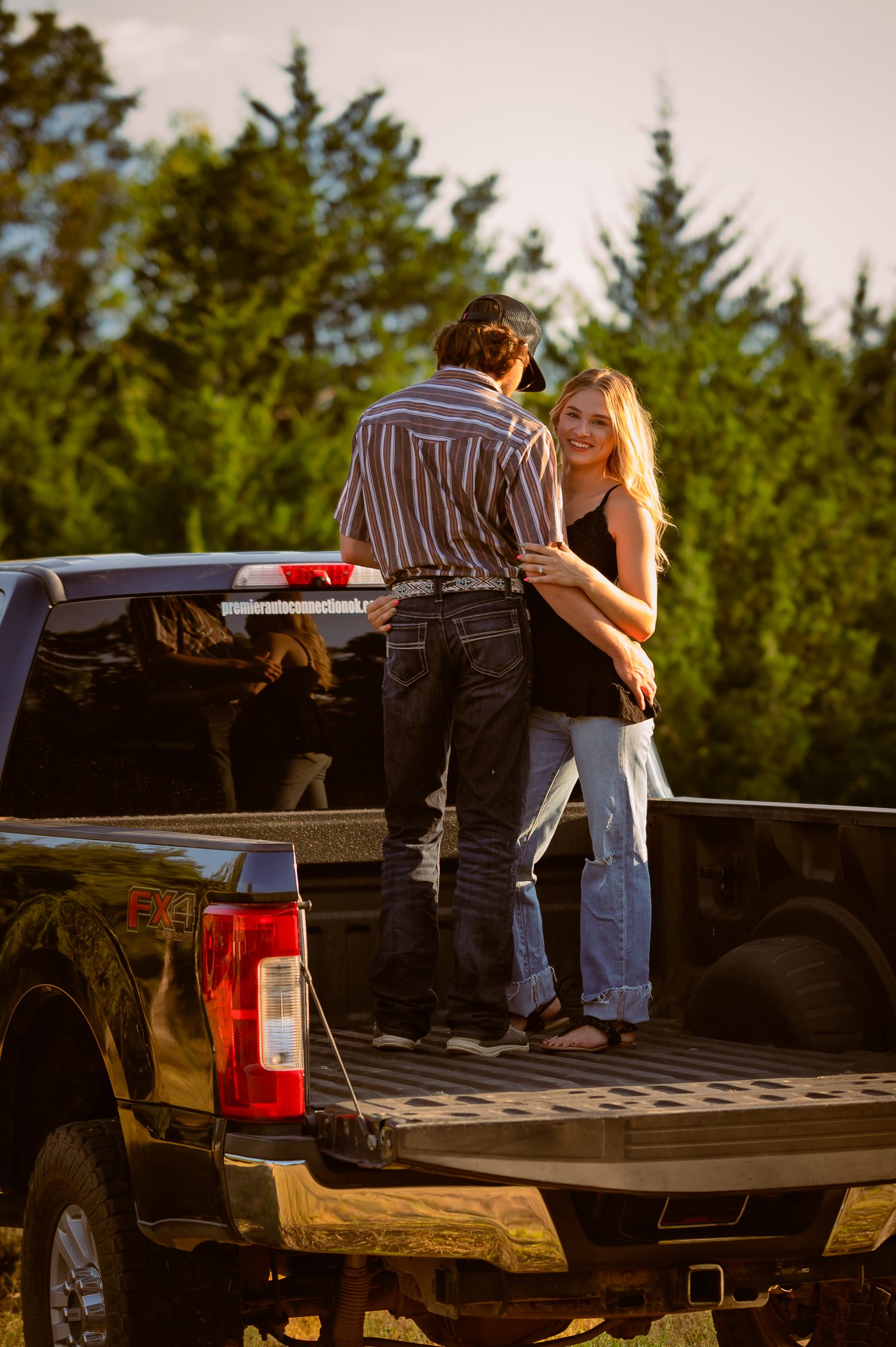 Couple standing in the bed of a black pickup truck surrounded by pine trees at sunset.