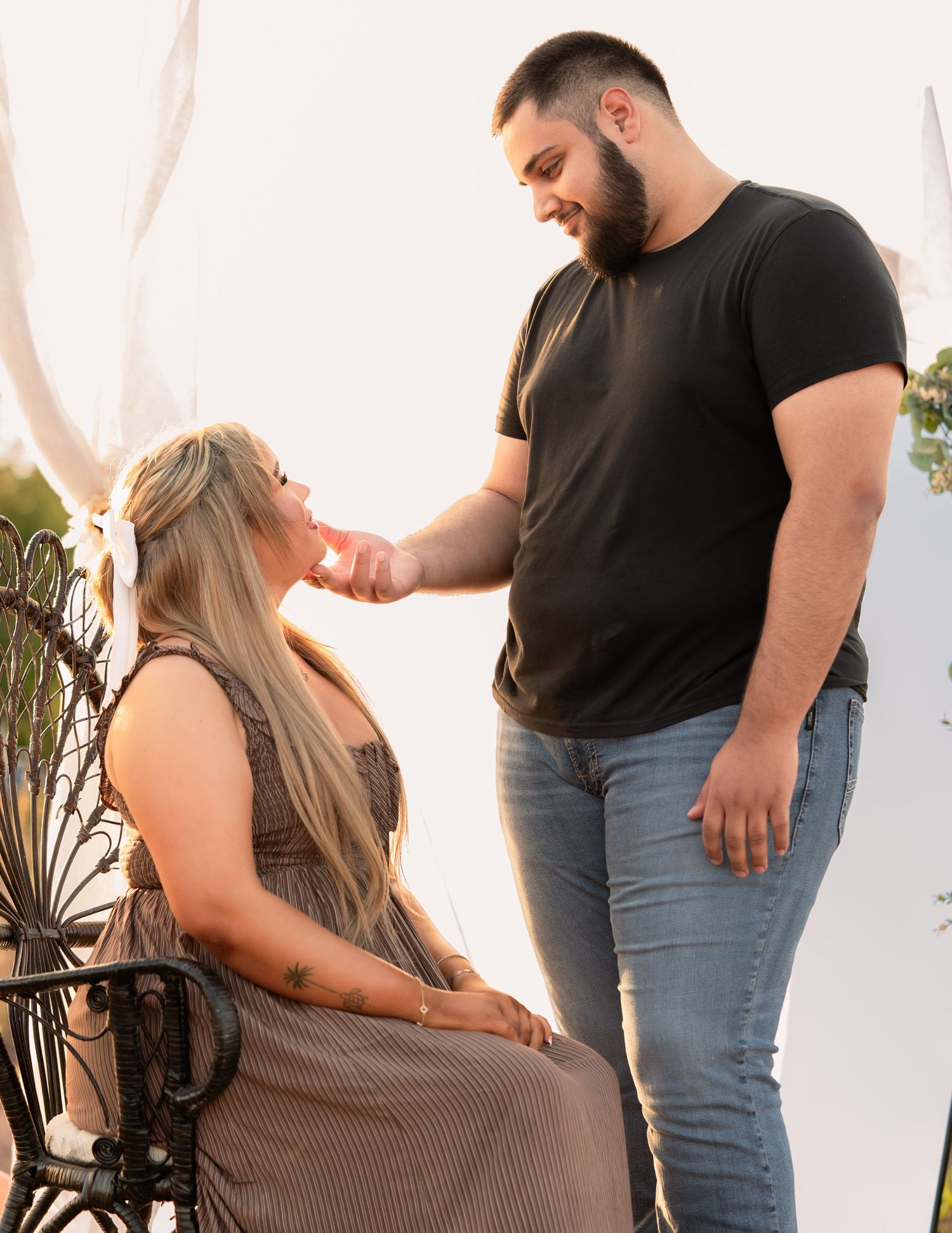 Man tenderly touching womans face as she sits in ornate chair during outdoor maternity shoot.