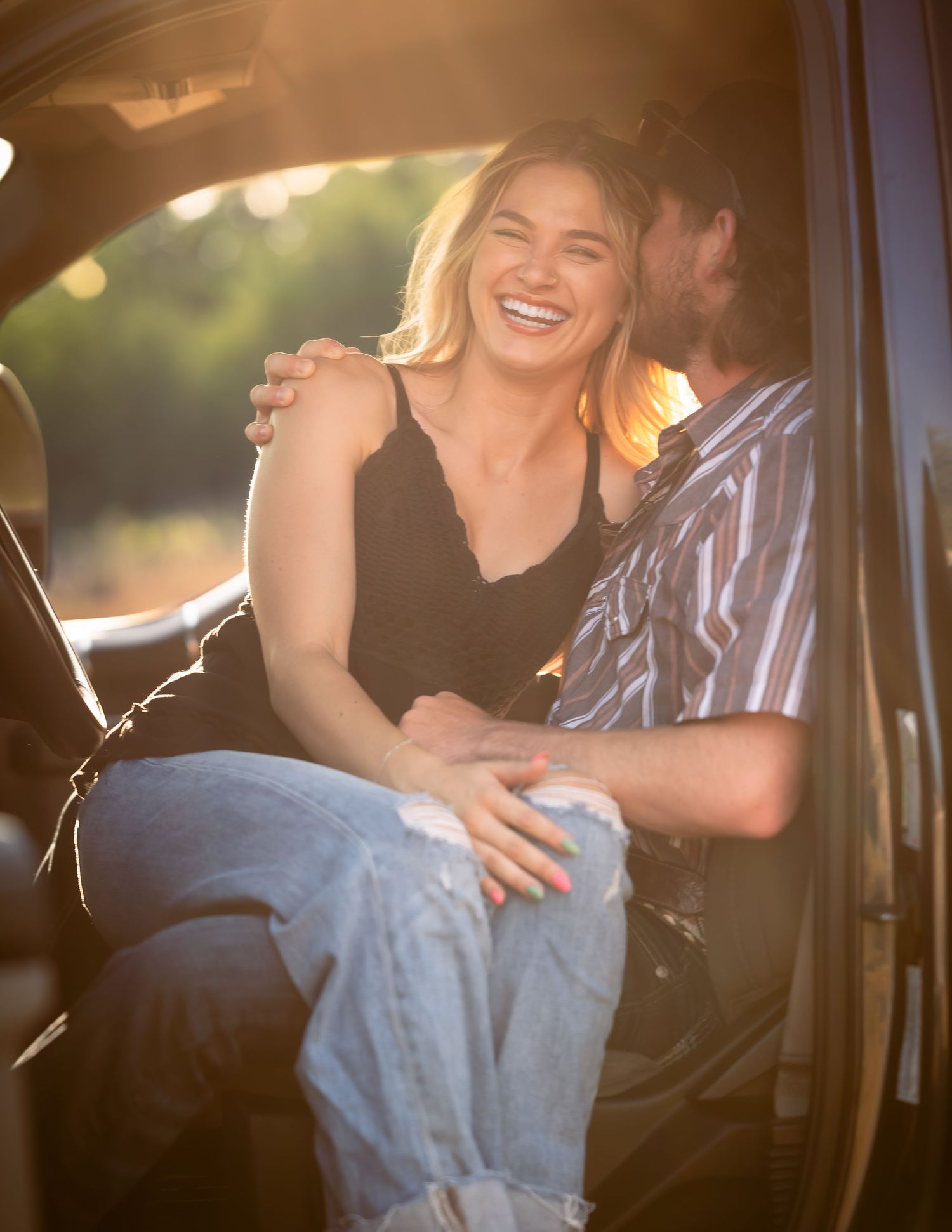 Laughing blonde woman sitting in truck cab with man during warm sunset engagement photos.