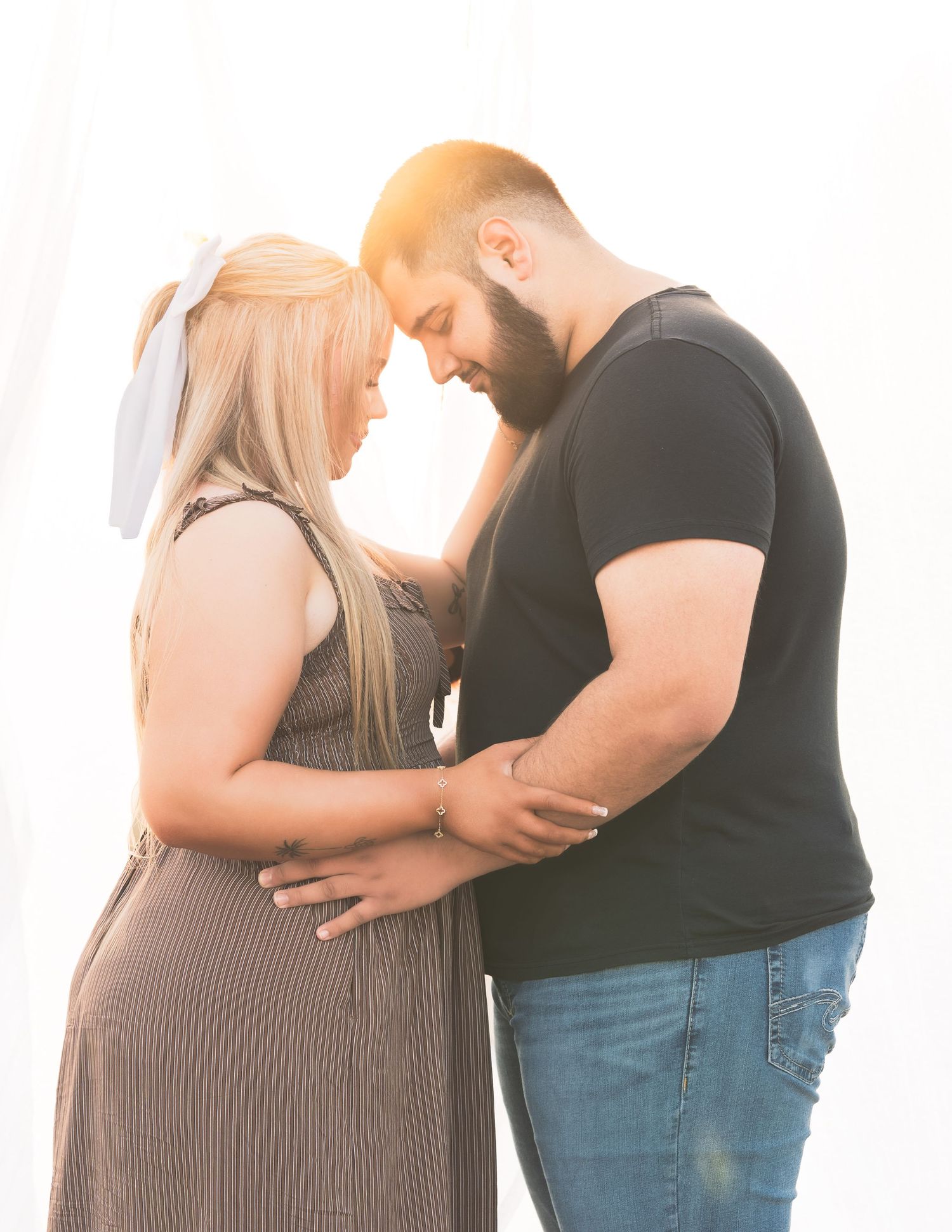 Expecting couple standing forehead to forehead, hands on baby bump, backlit by golden sunset light.