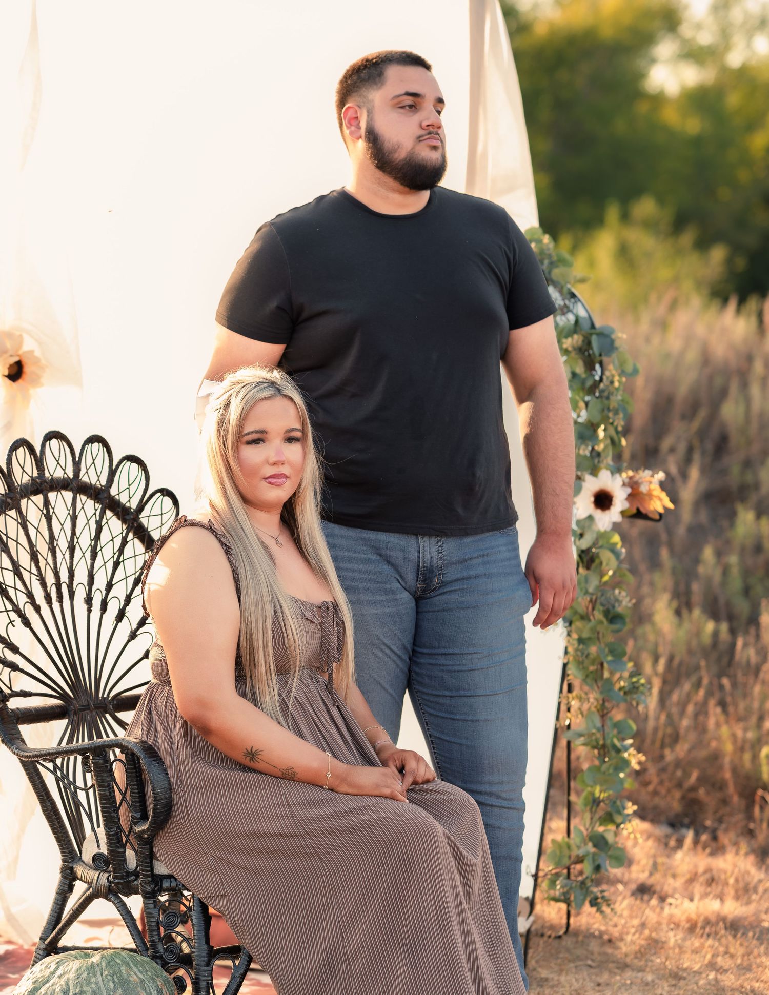 Couple posing outdoors at golden hour, woman seated in ornate chair, man standing behind her.