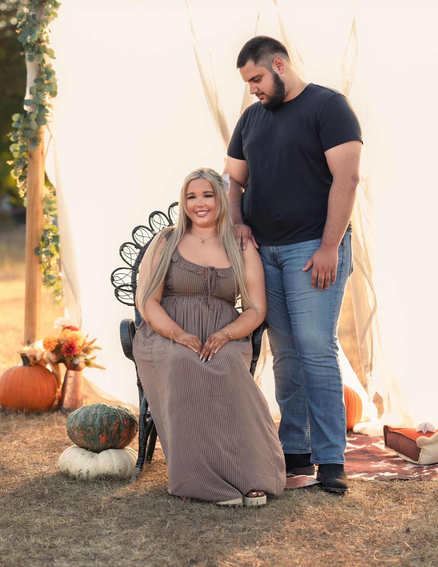Pregnant woman in maxi dress seated in ornate chair with partner standing beside her outdoors.