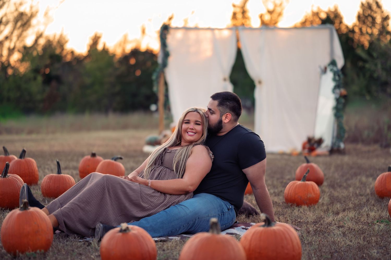 Man kissing smiling woman on cheek while sitting in pumpkin patch at golden hour.
