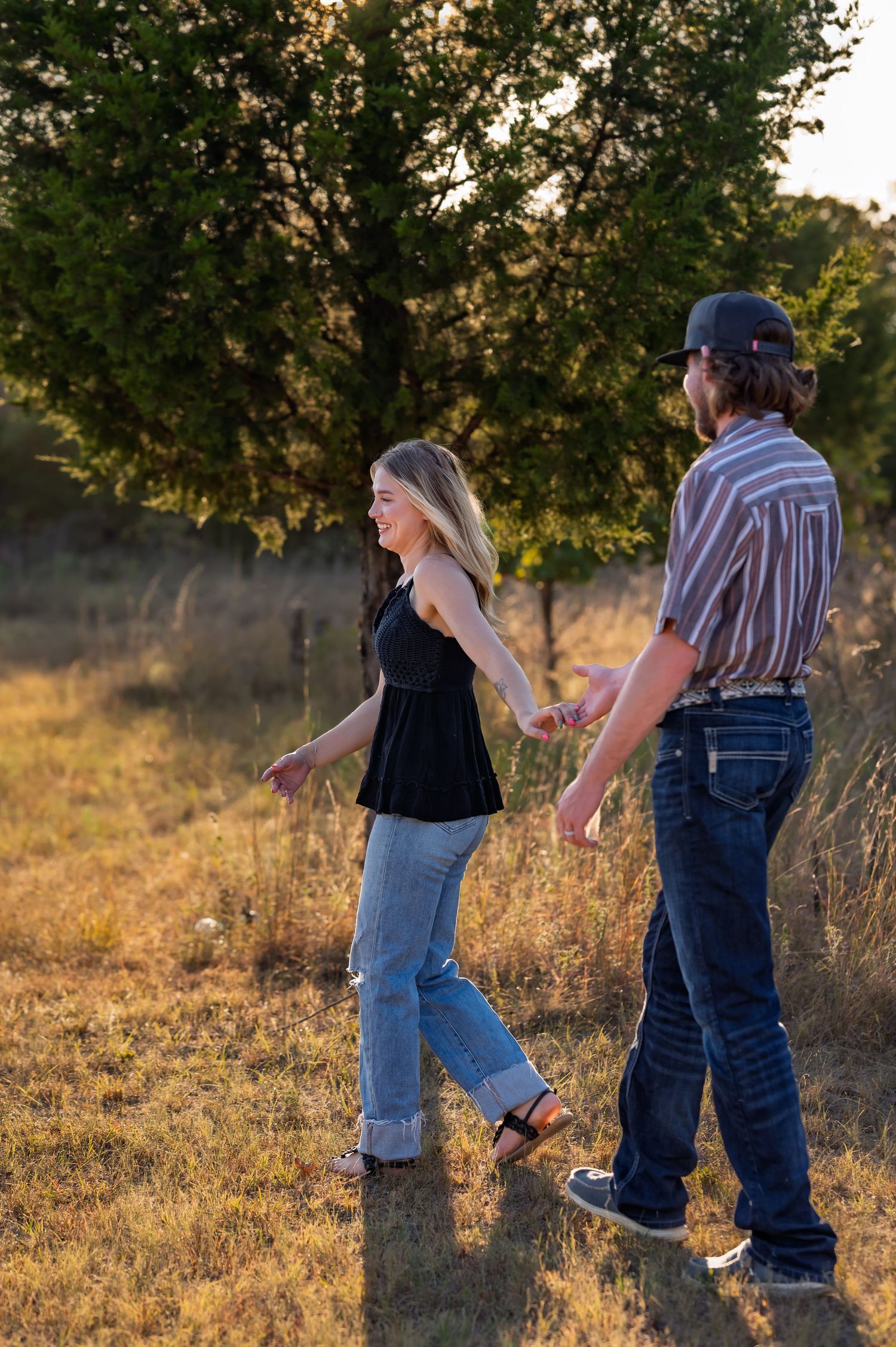 Happy couple holding hands and walking through a golden meadow with trees at sunset.