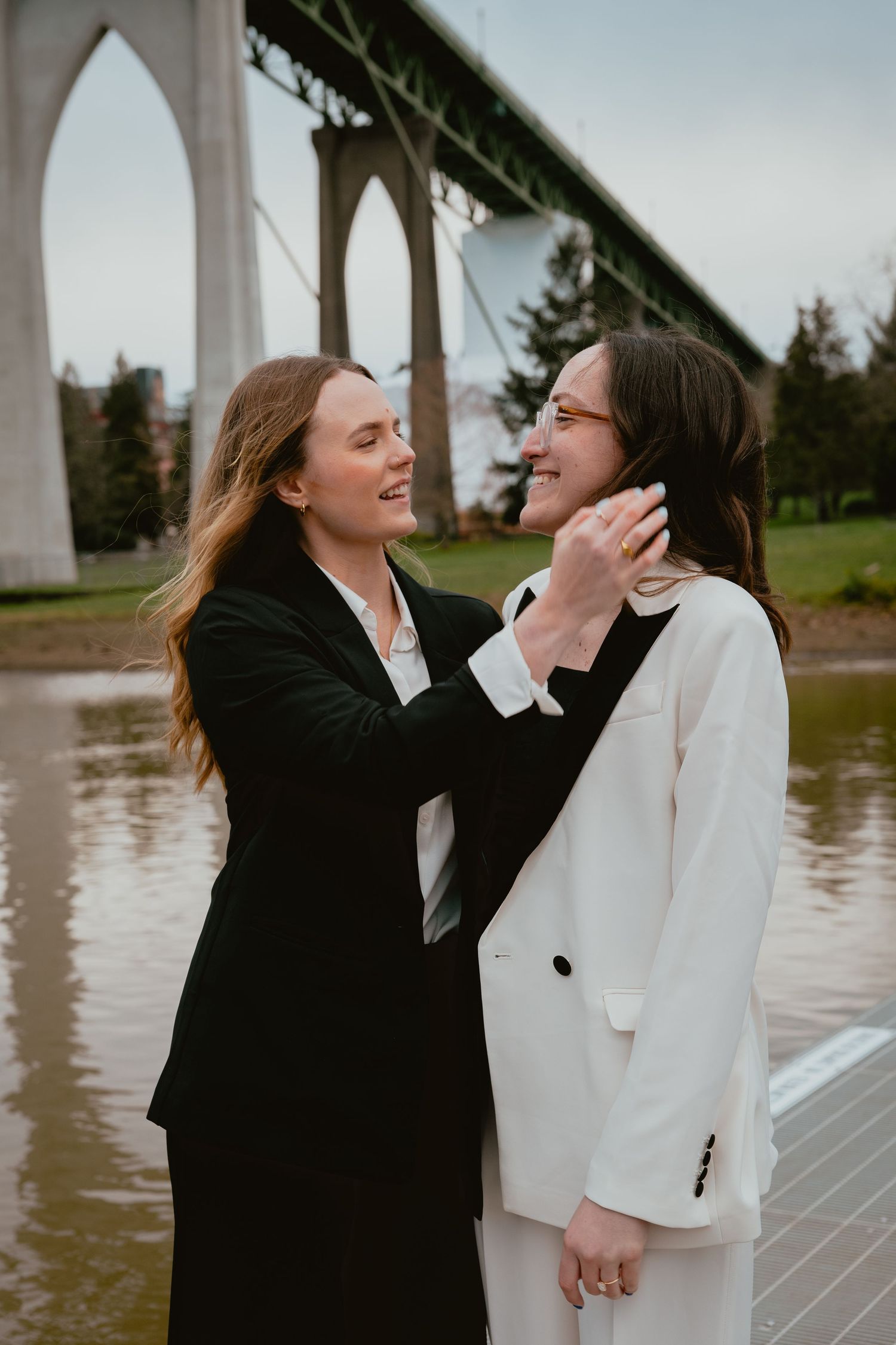 Two women in black and white suits sharing a tender moment near a bridge.