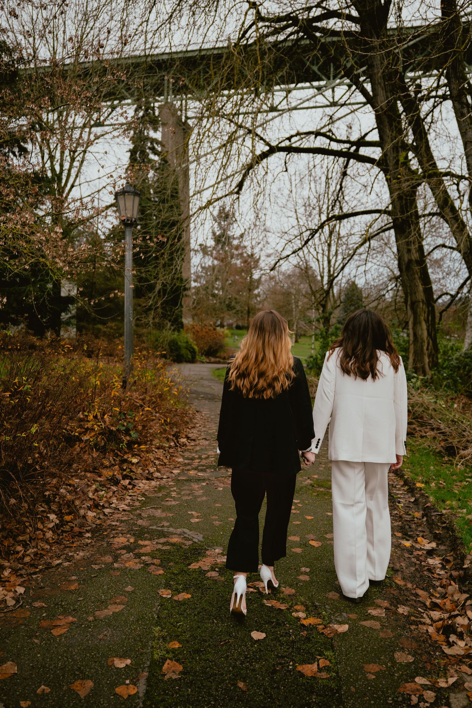 Two women walk hand in hand along an autumn park path under bare trees.