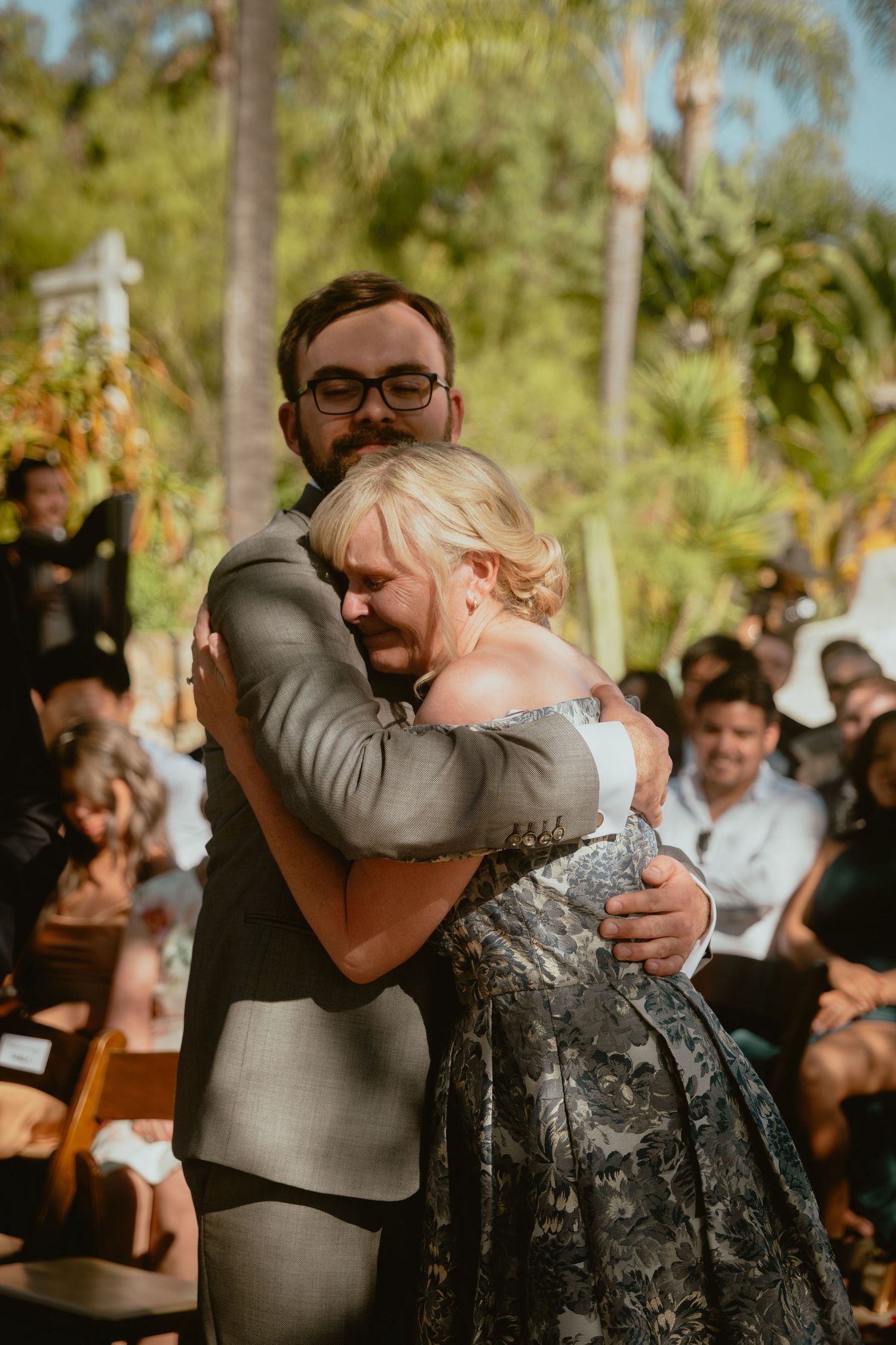 Emotional groom in gray suit shares a heartfelt hug with a woman during an outdoor wedding ceremony.