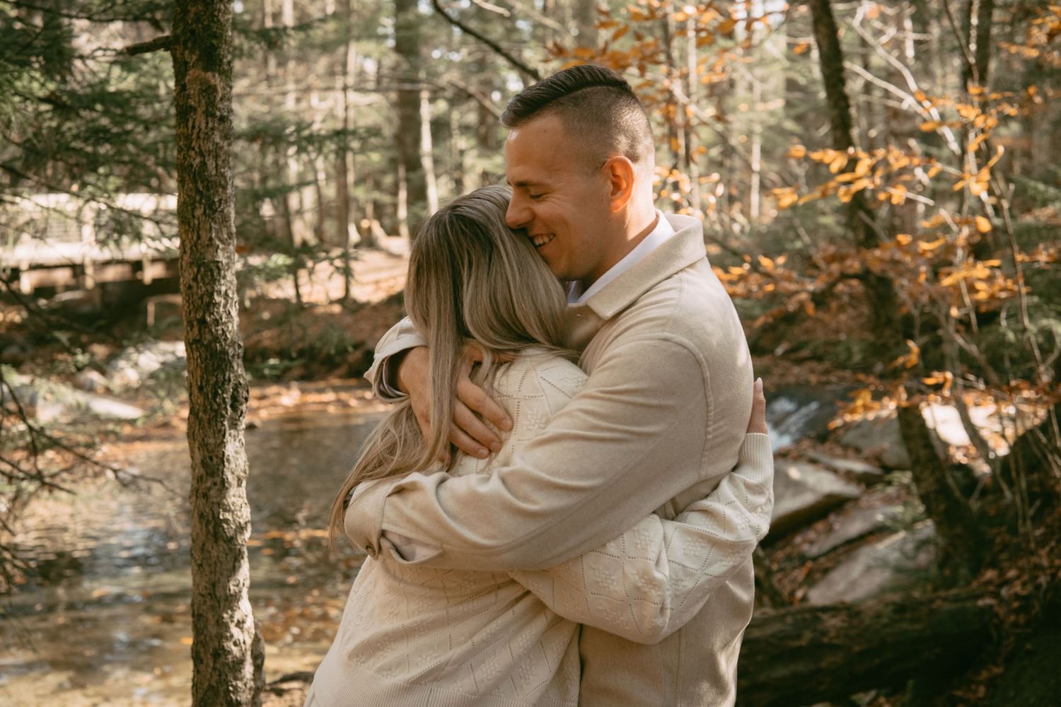 Couple shares a warm embrace in a sunlit autumn forest near a stream.