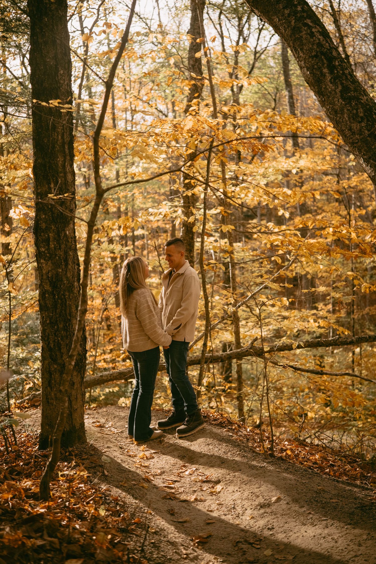 Couple holding hands on a sunlit autumn forest trail surrounded by golden fall foliage.