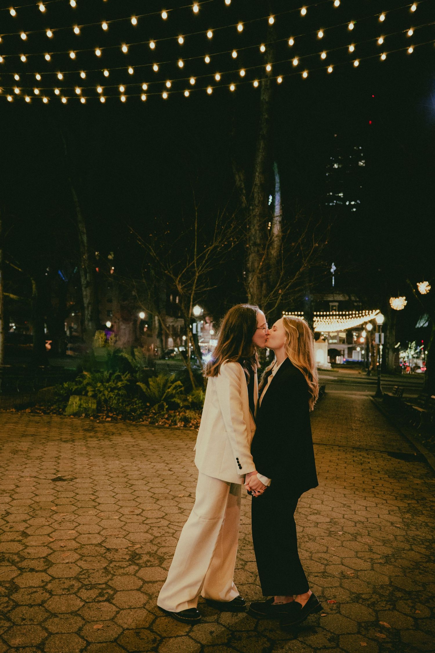 Same-sex couple kisses under string lights in a park at night, both wearing suits.