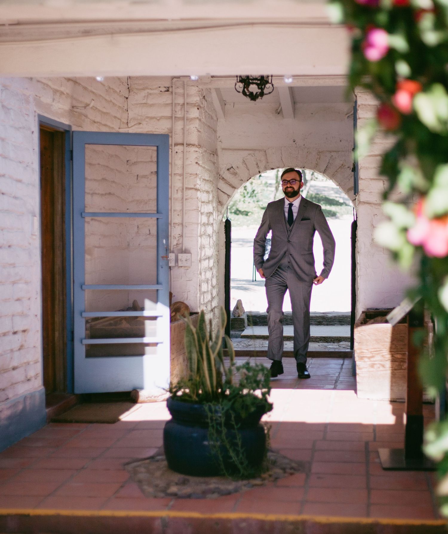 Dapper groom in grey suit stands in a rustic archway entrance of a charming wedding venue.