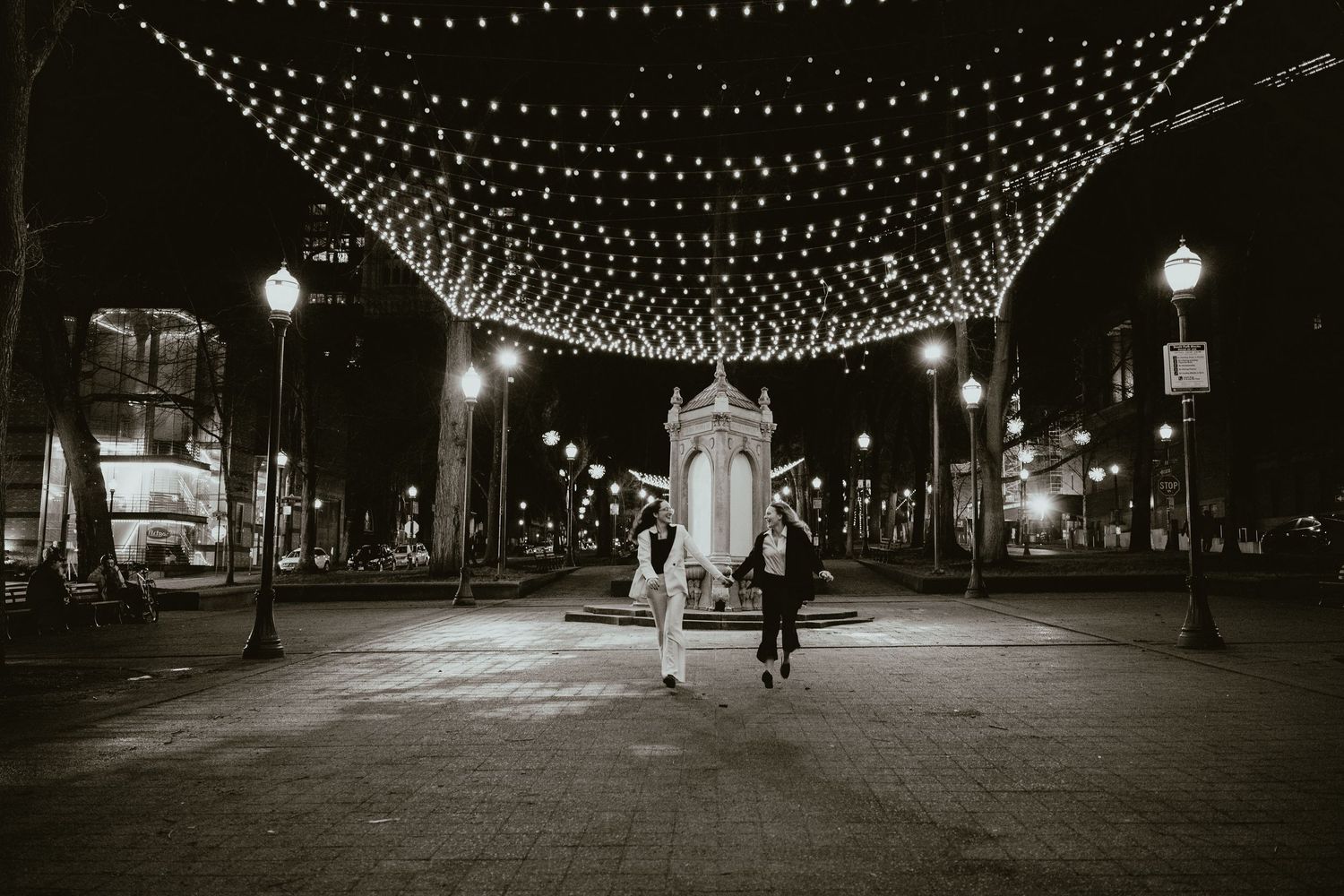 Two women dance under canopy string lights in a wide black and white nighttime city plaza.