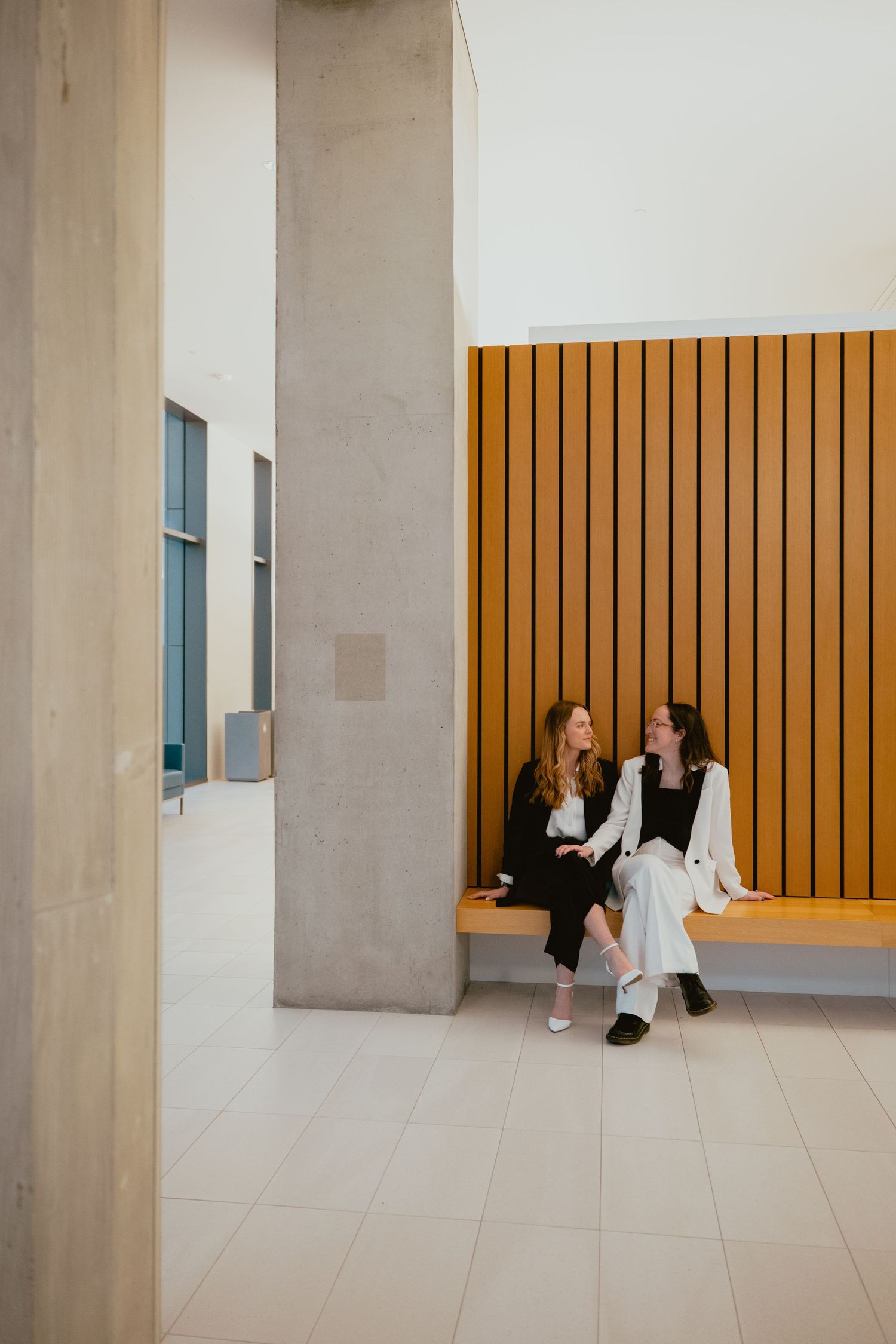 Two women sit together on a wooden bench in a modern building interior with warm wood panel walls.