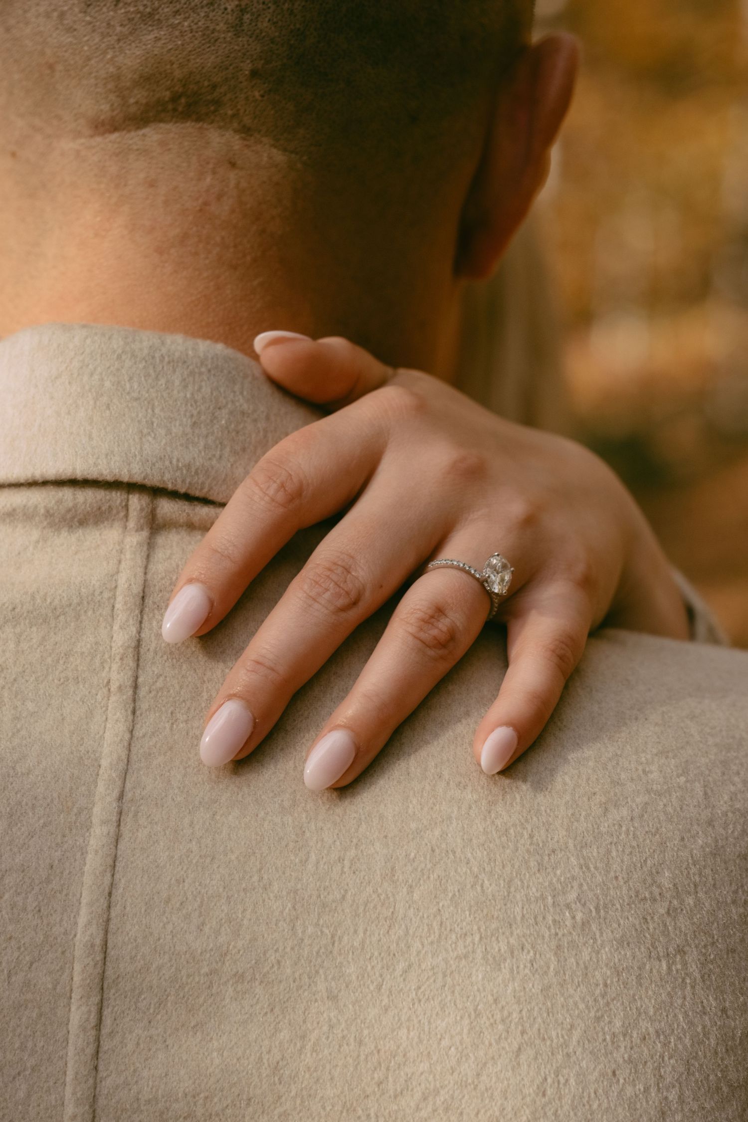 Close-up of a woman's hand with a diamond engagement ring resting on a man's shoulder.