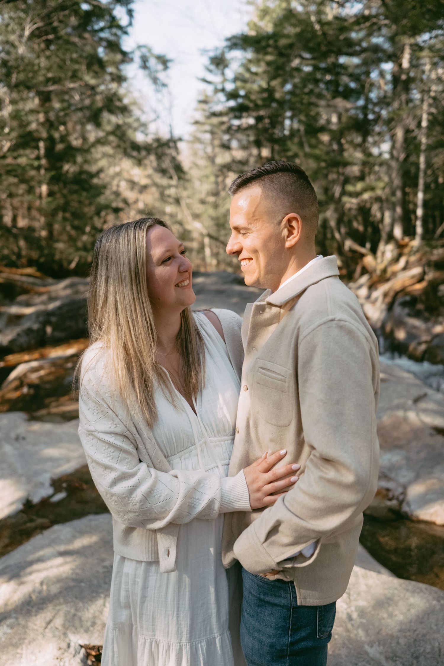 Couple smiles at each other while embracing on rocky terrain surrounded by pine trees.