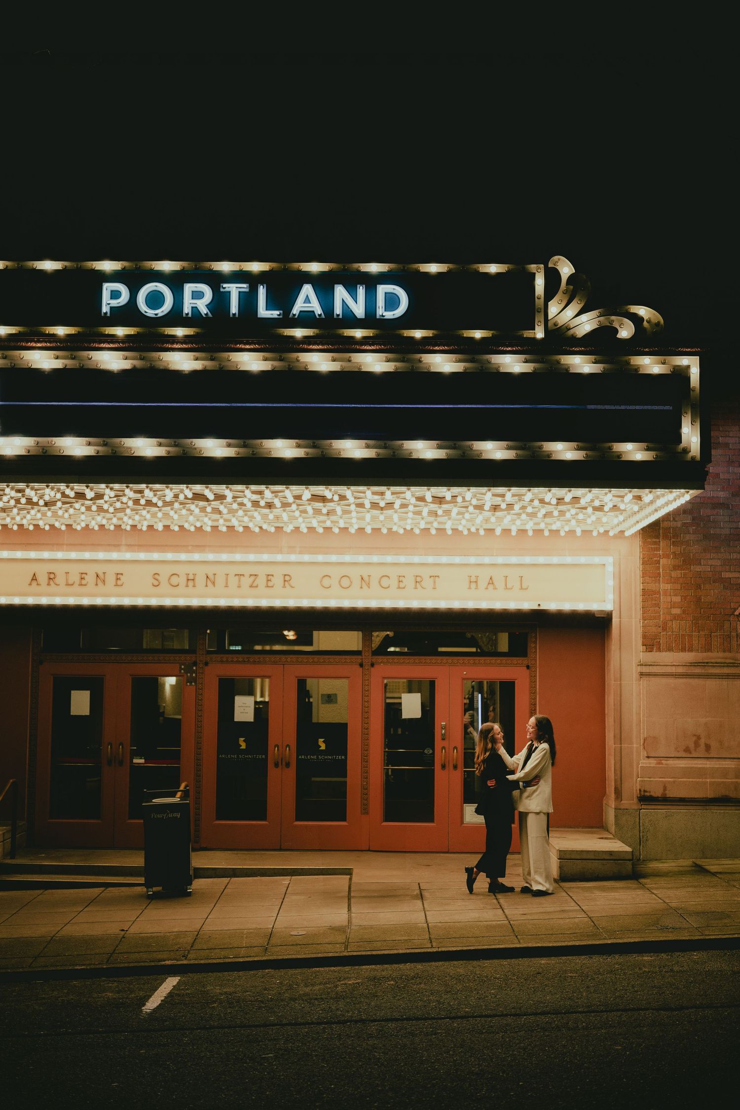 Couple embraces under the lit marquee of Arlene Schnitzer Concert Hall in Portland at night.