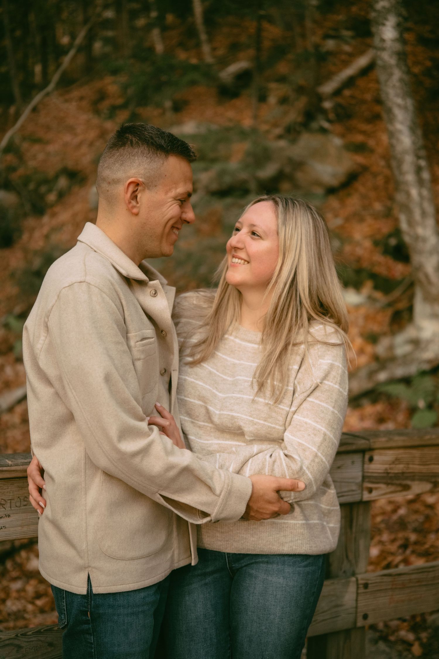 Happy couple embracing and smiling at each other by a wooden fence in fall forest.