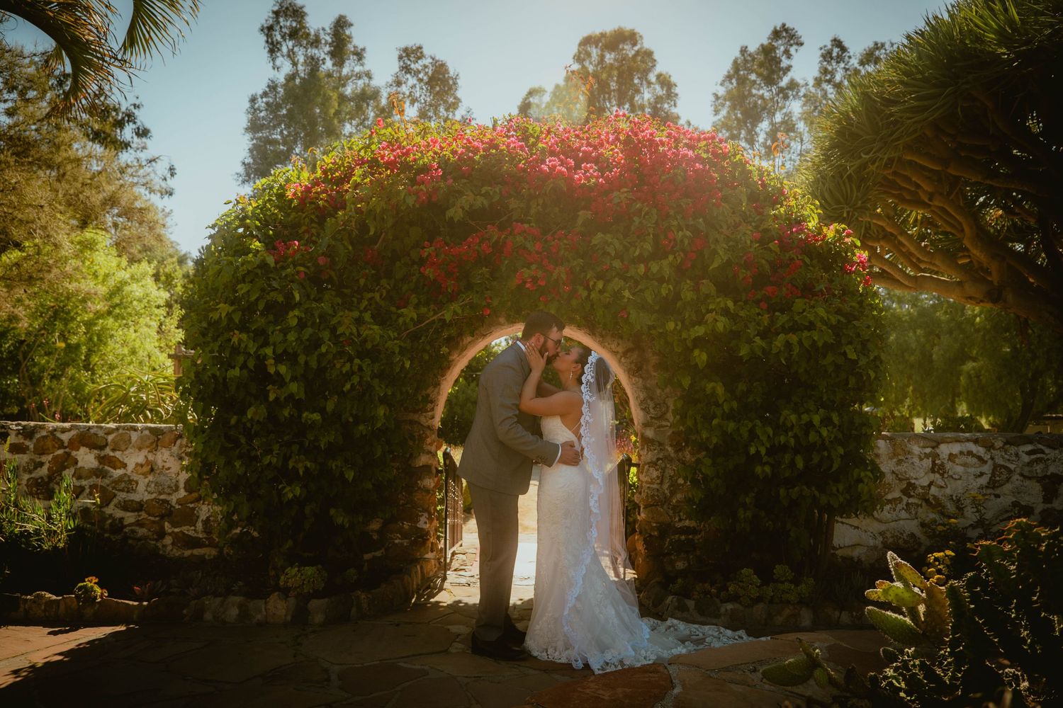 Bride and groom share a romantic kiss under a floral arch covered in red bougainvillea at an outdoor wedding.