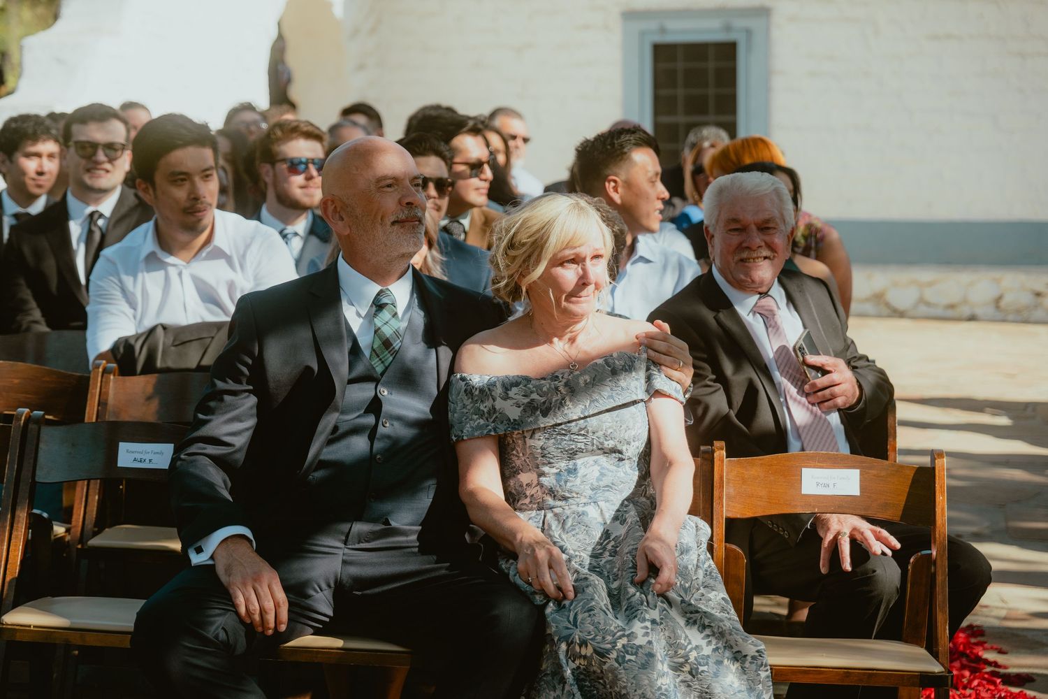 Wedding guests seated in wooden pews at an outdoor ceremony, smiling couple in foreground dressed in formal attire.