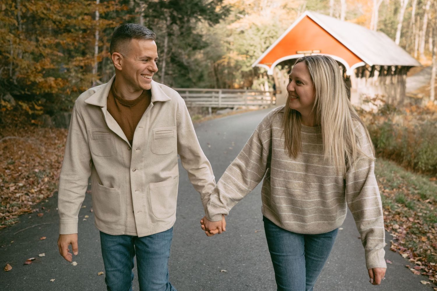 Couple walks hand in hand on a road with a red covered bridge in fall foliage.