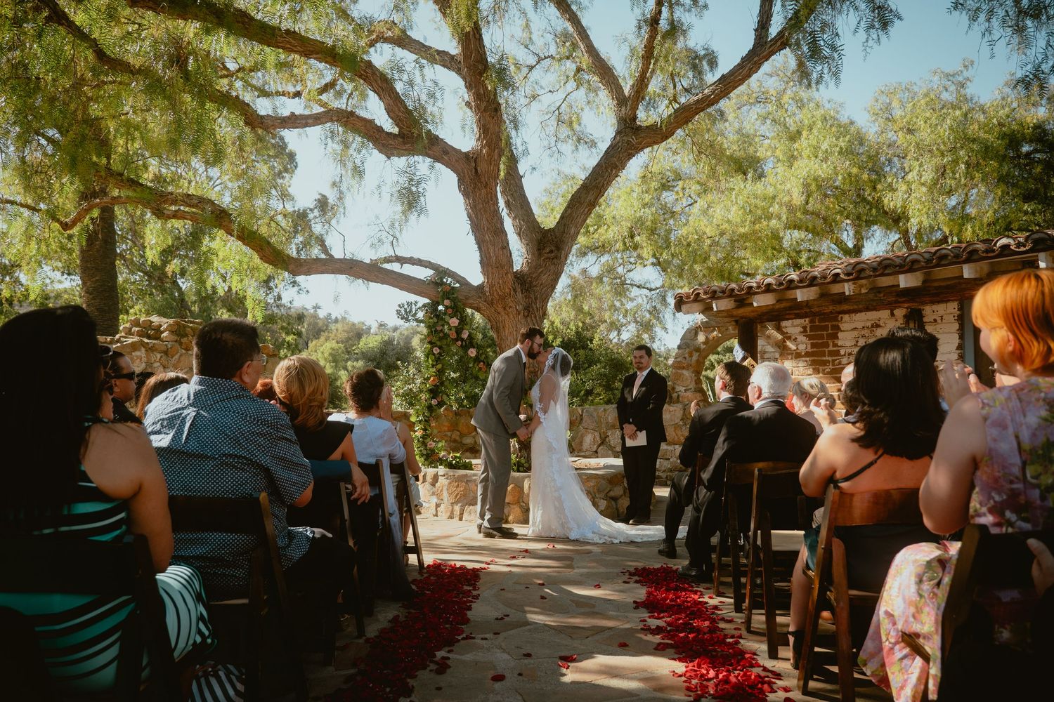 Wedding guests watch as bride and groom share a kiss at an outdoor ceremony.