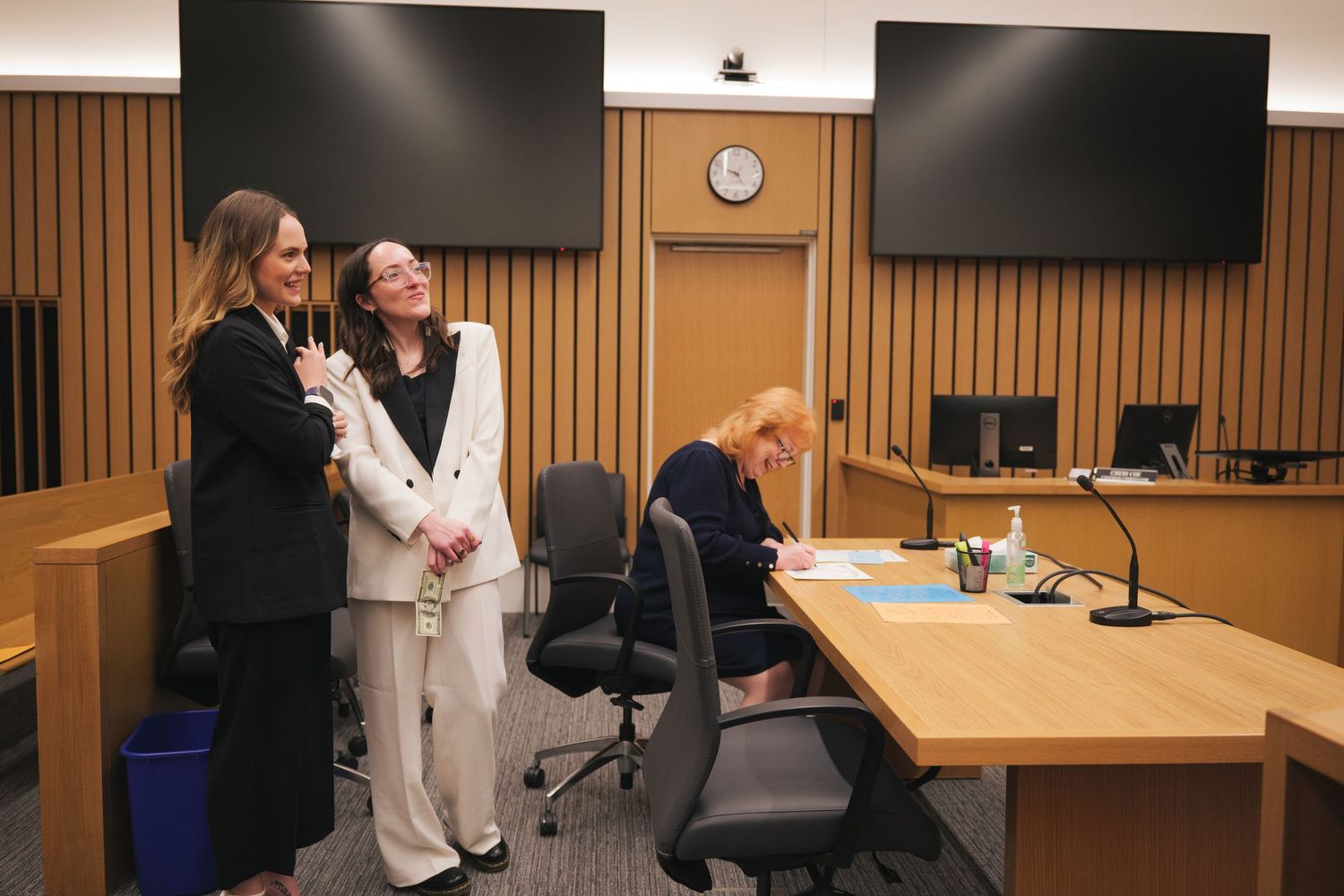 Two women in formal attire smile together inside a courtroom with wood-paneled walls.