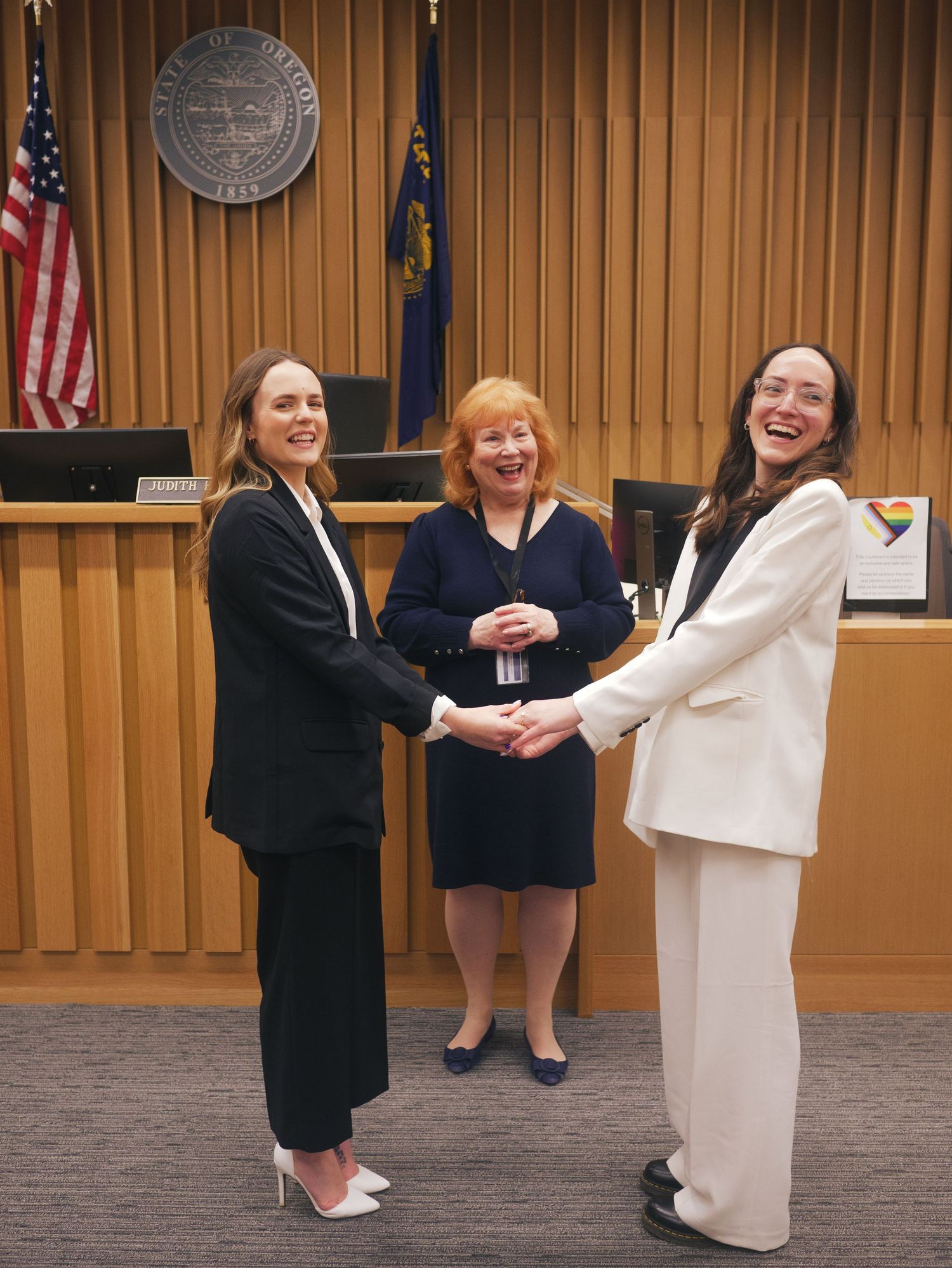 Two women in formal attire shake hands in a courtroom while a judge smiles between them.