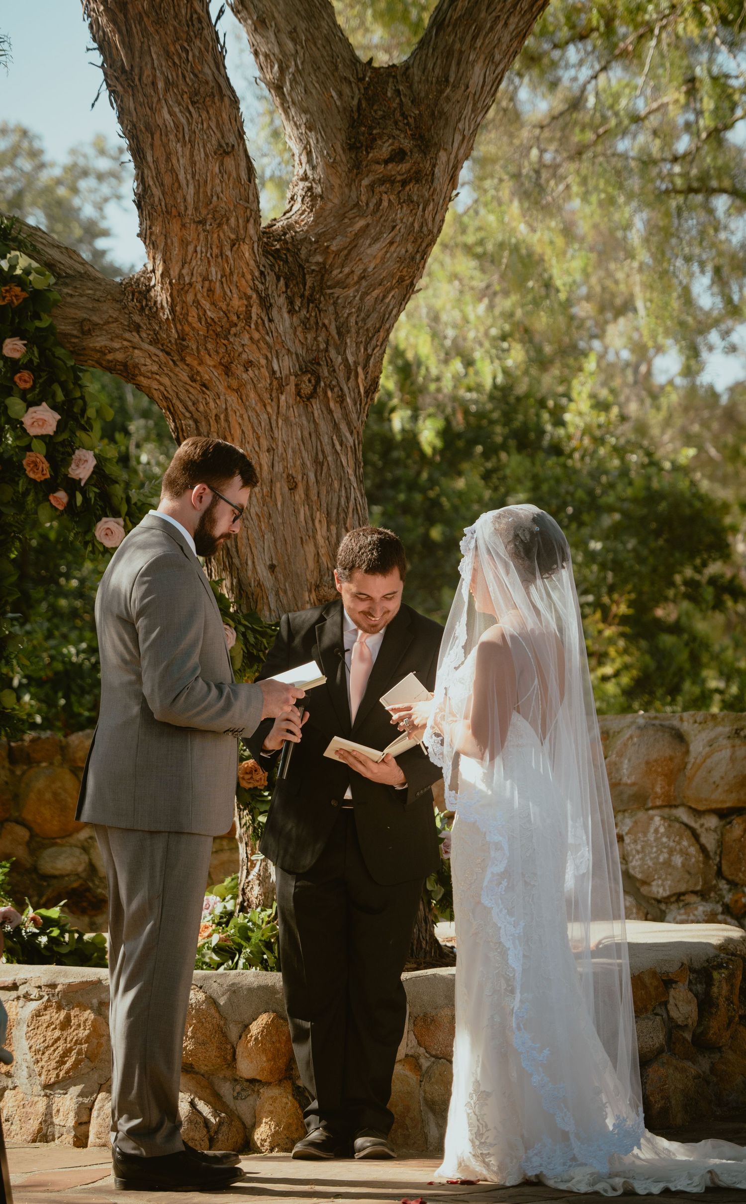 Bride and groom exchange vows with officiant under a large tree at an outdoor ceremony.