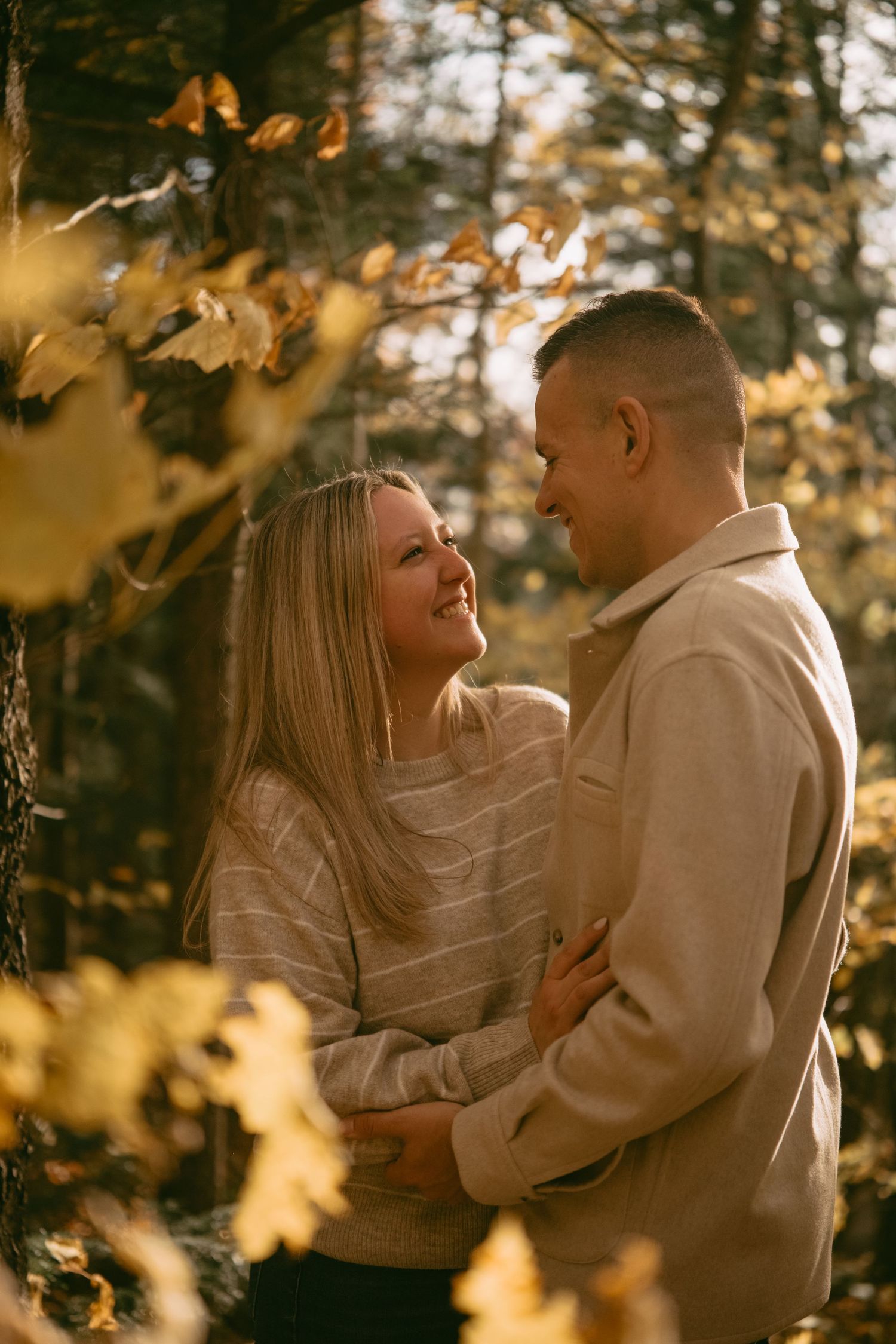 Happy couple hugging and smiling at each other in a sunlit fall forest setting.