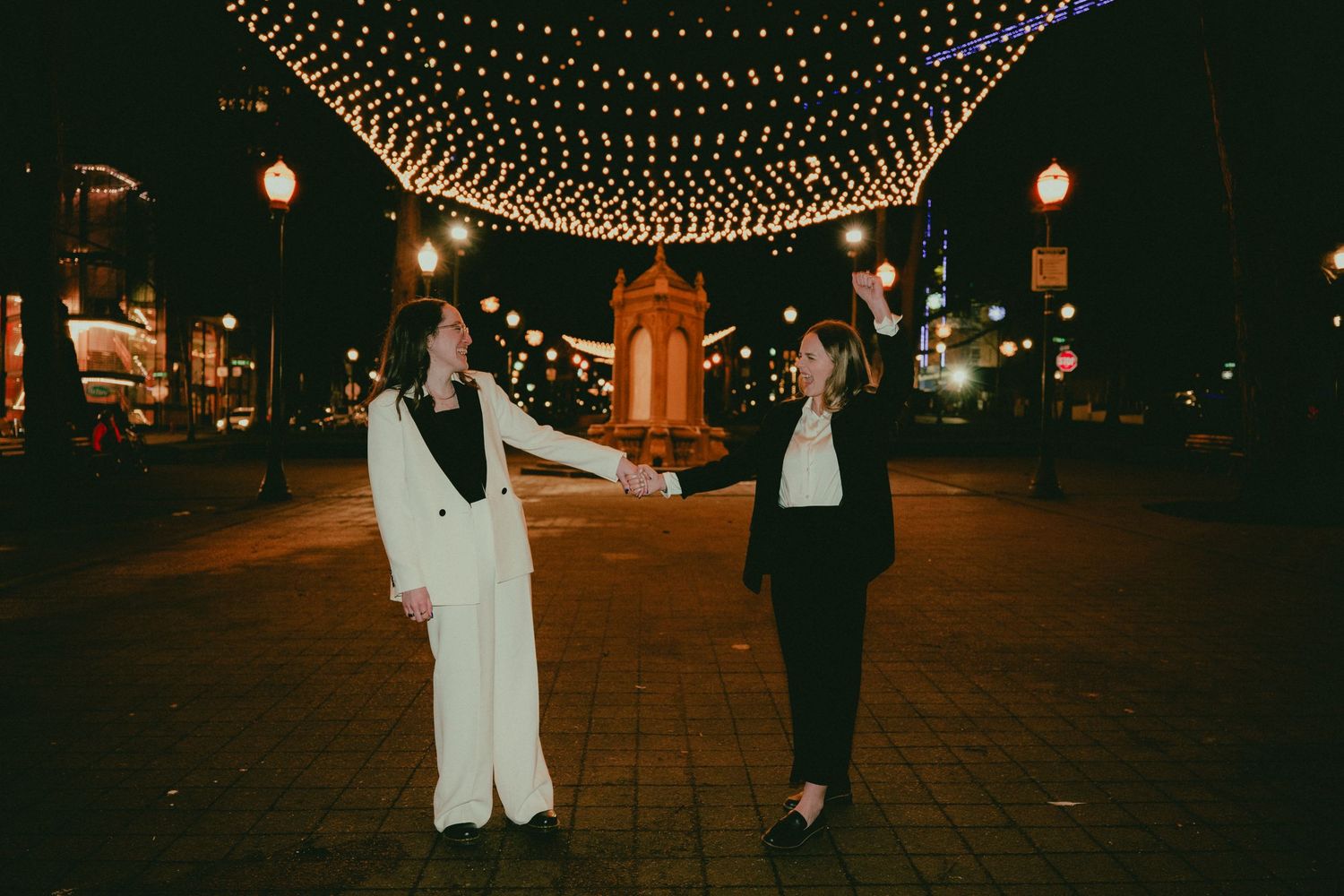 Two women in suits hold hands on a city street lit by overhead string lights at night.