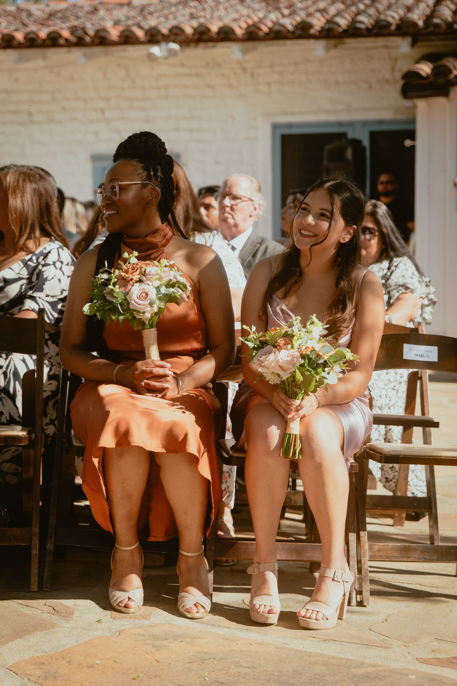 Two bridesmaids holding bouquets and smiling at sunny outdoor wedding ceremony.