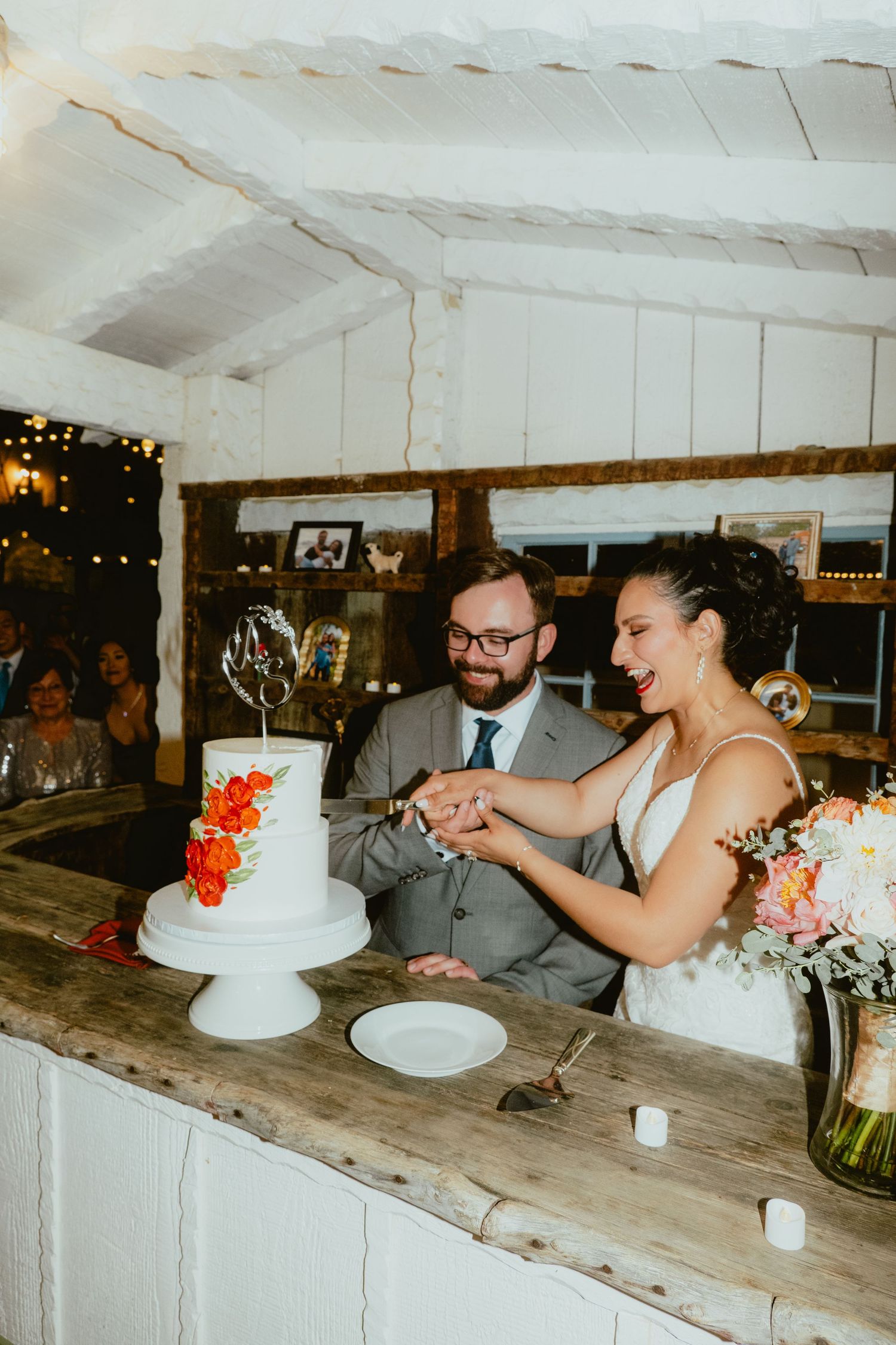 Smiling newlyweds cut their white two-tier wedding cake decorated with orange flowers at reception.