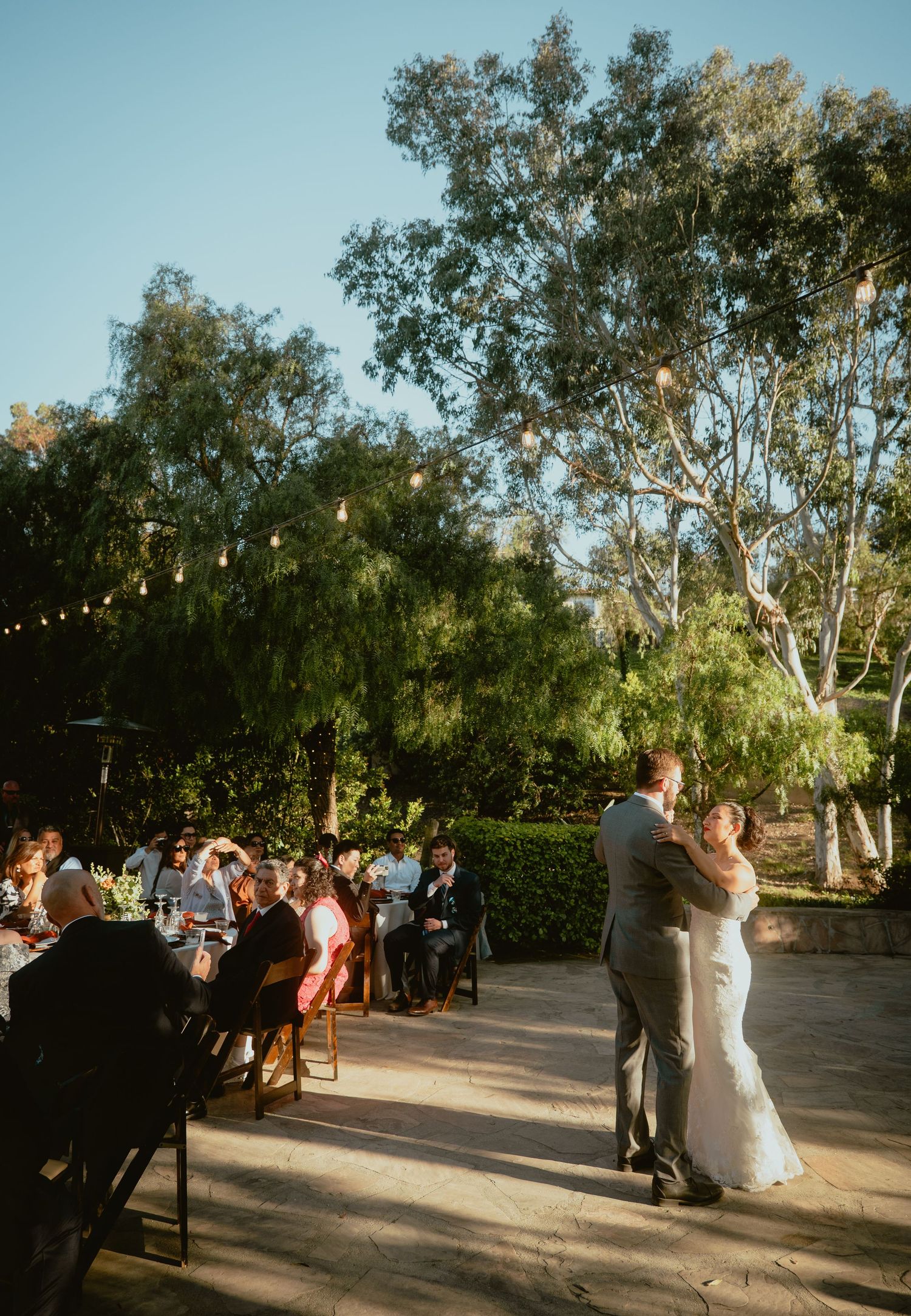 Newlyweds share their first dance at an outdoor wedding reception with string lights.