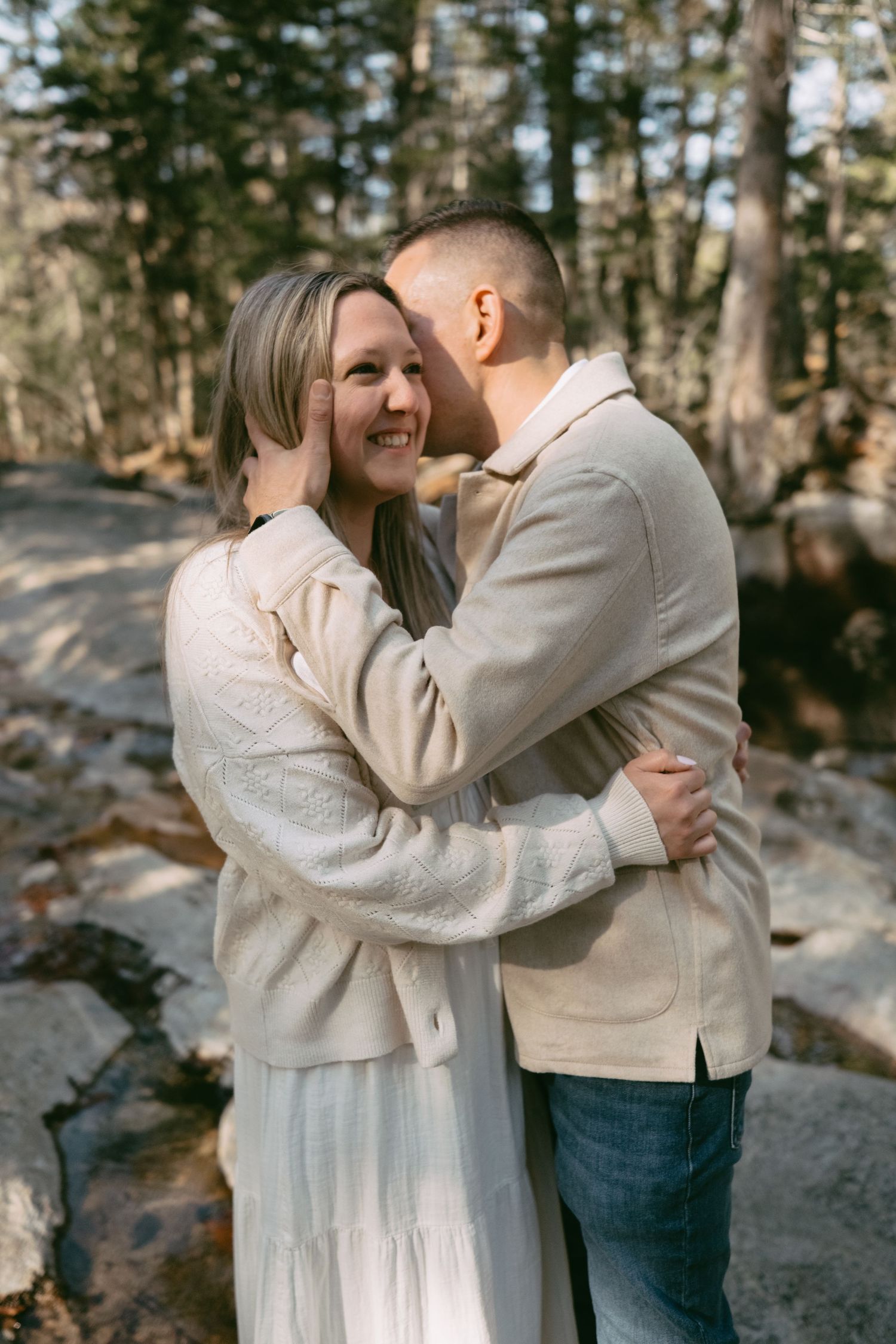 Smiling woman in white dress embraced by man in beige jacket in a forest setting.