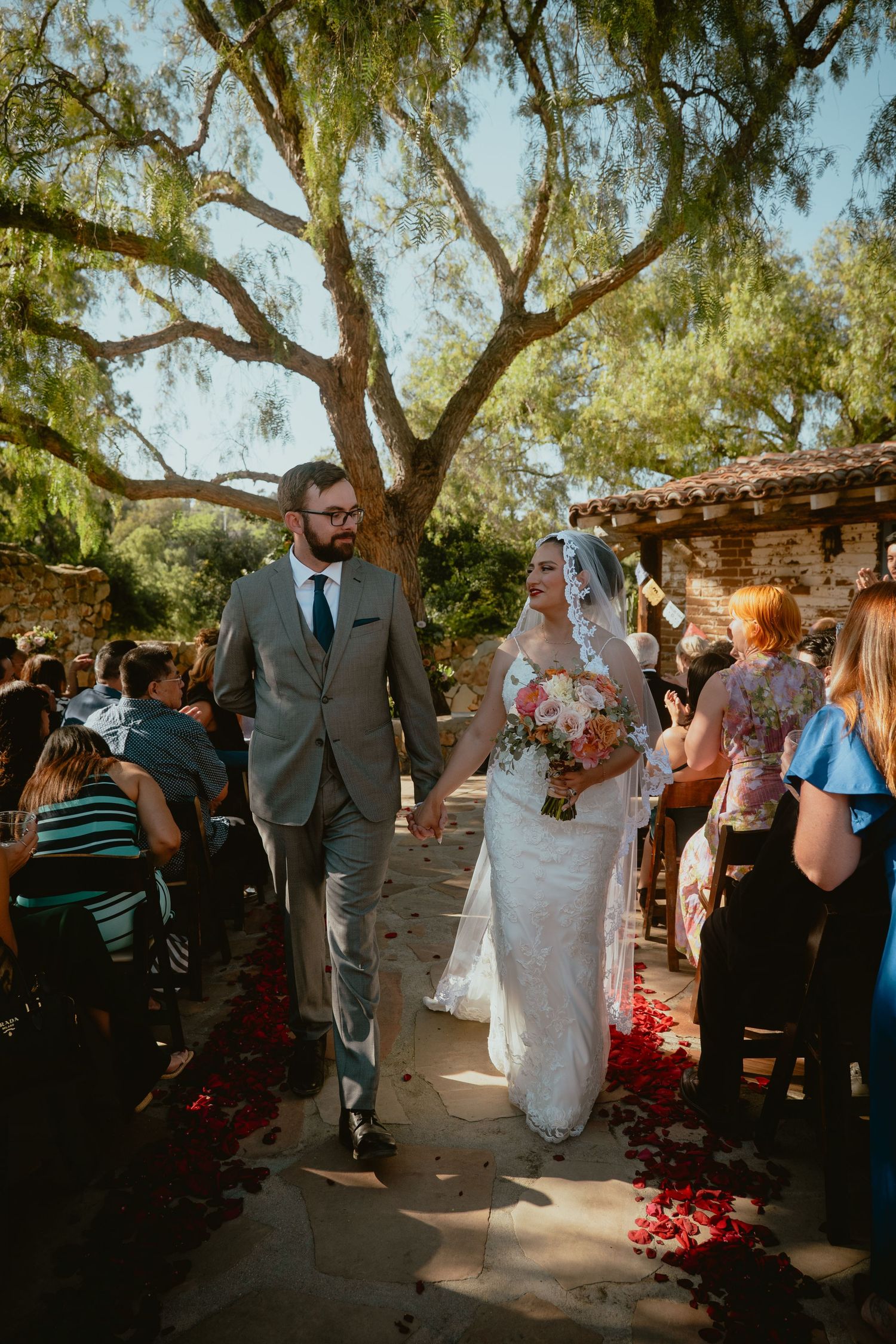 Newlyweds walk down petal-lined aisle after outdoor wedding ceremony under large tree.