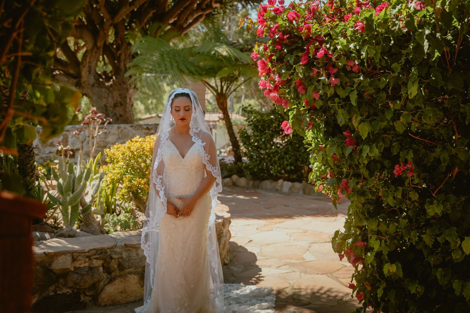 Elegant bride in lace wedding gown and veil stands in a sunlit garden with vibrant pink bougainvillea.