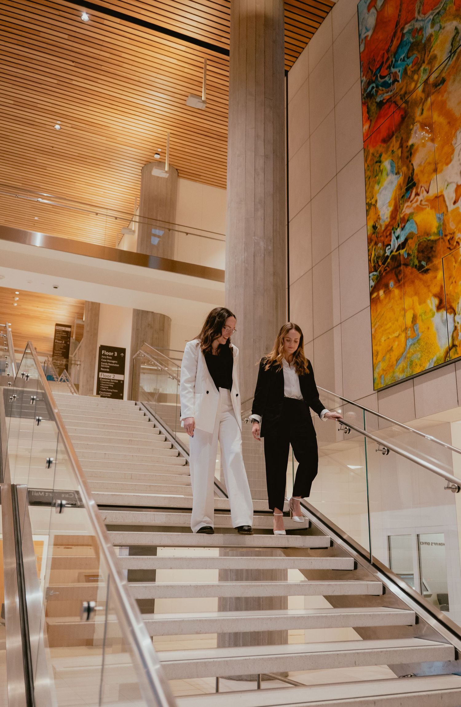 Two women in black and white suits walking down modern interior stairs together.