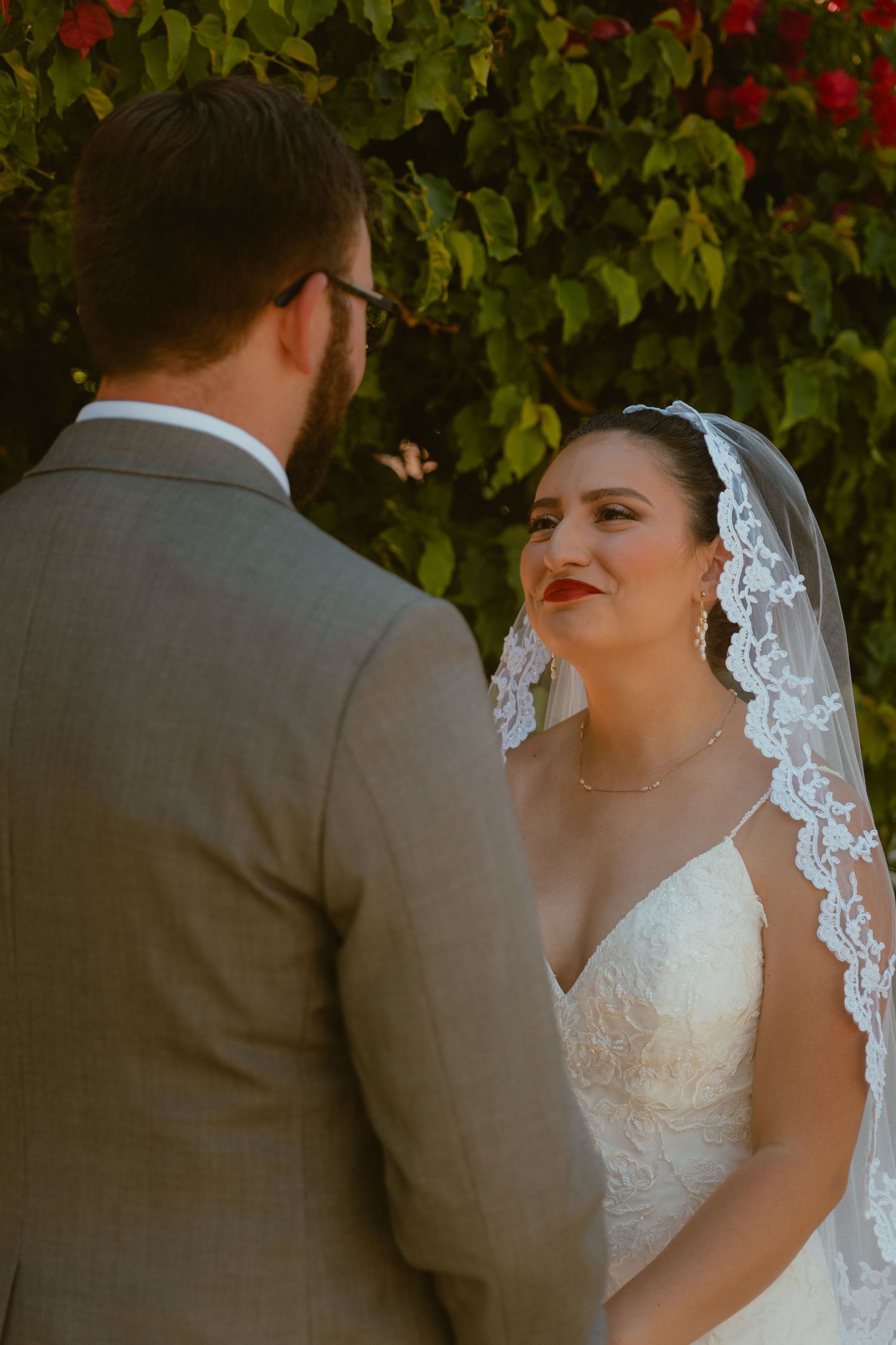 Bride gazes lovingly at groom wearing a lace veil at their outdoor wedding ceremony.