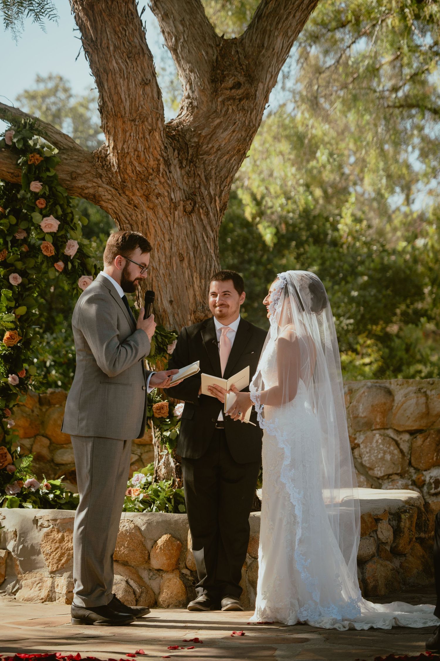 Groom reads vows to bride before officiant at an outdoor wedding ceremony with stone wall backdrop.