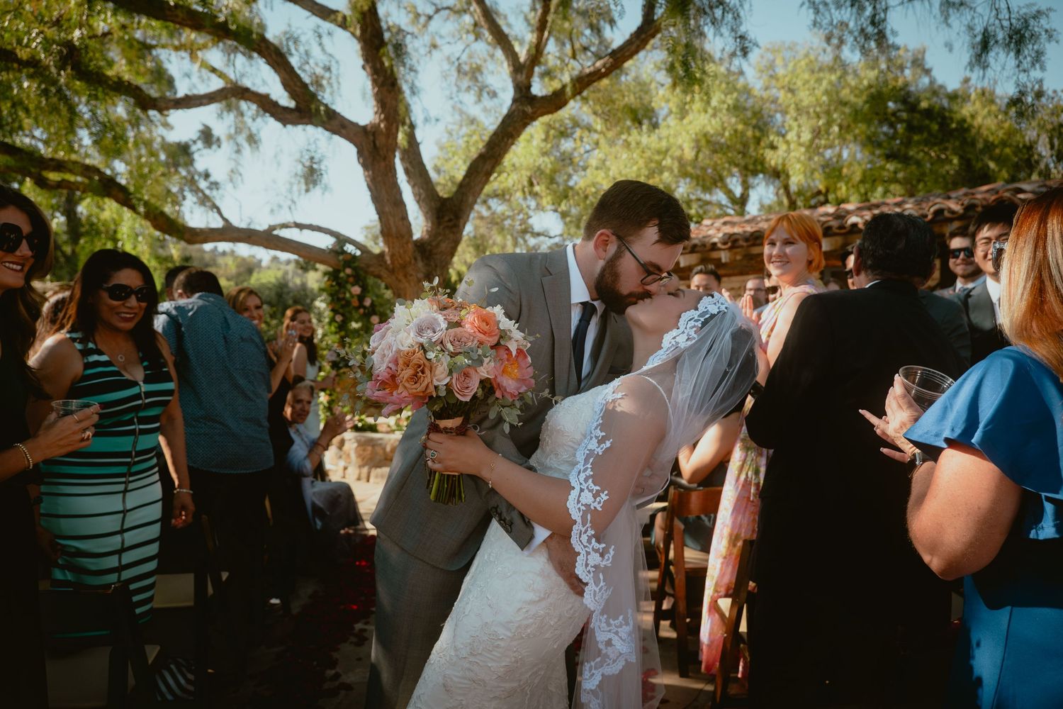 Bride and groom share a passionate kiss surrounded by cheering guests after their outdoor wedding ceremony.