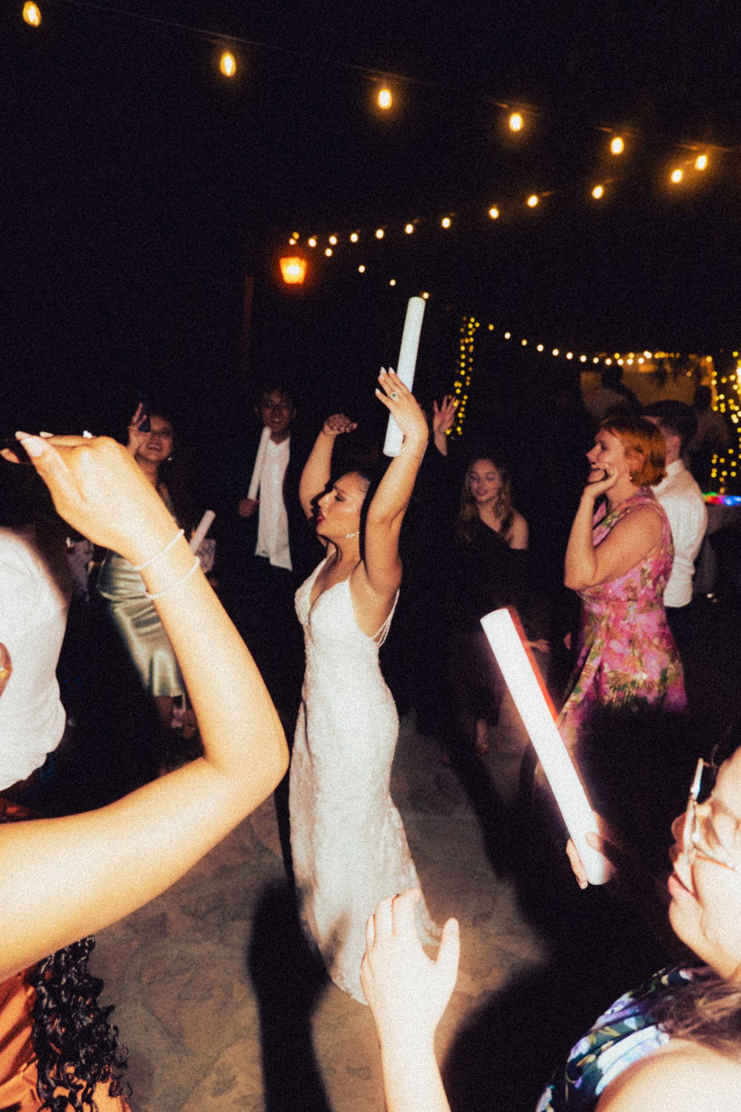 Bride in white gown dancing with glow sticks at an outdoor wedding reception under string lights.