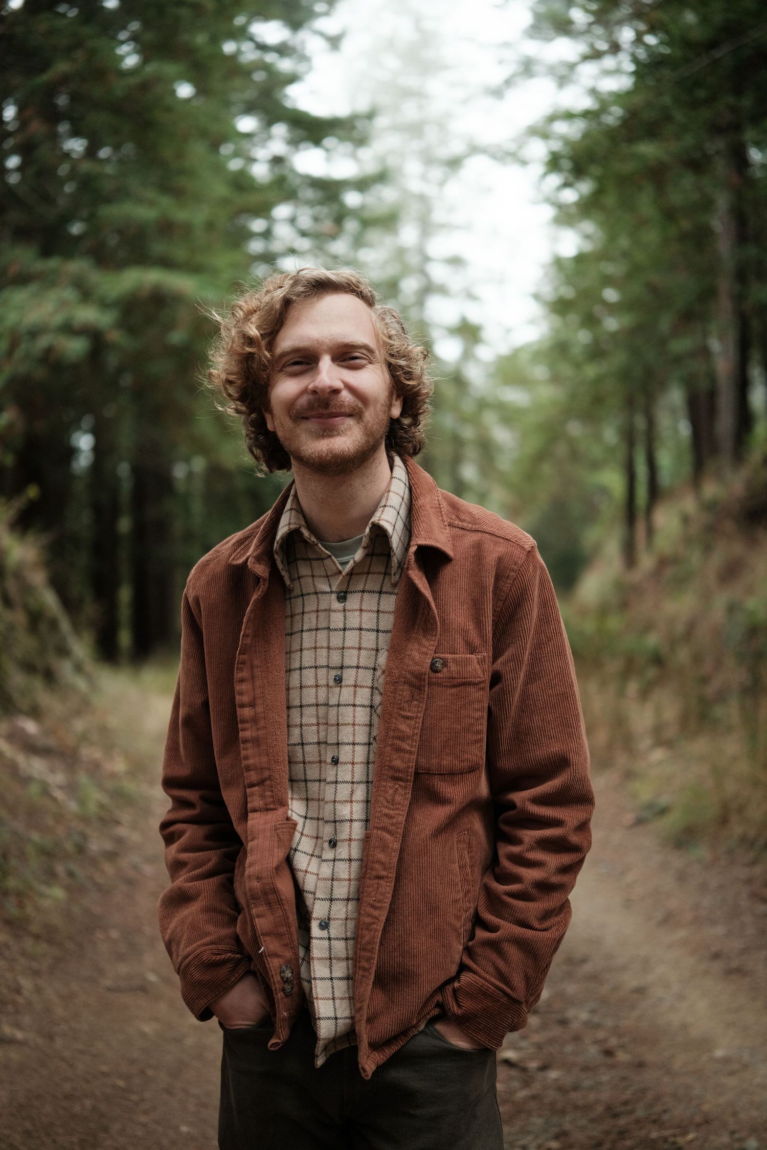 Smiling young man with curly hair standing on a forest dirt path wearing a brown jacket.