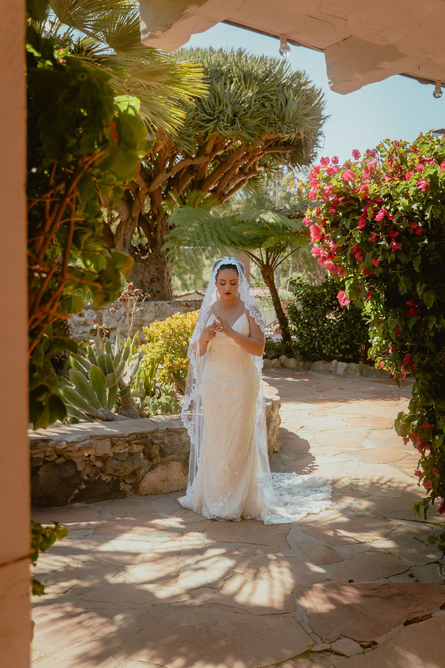 Bride in elegant white lace gown and veil standing in a sunlit garden pathway surrounded by tropical flowers.