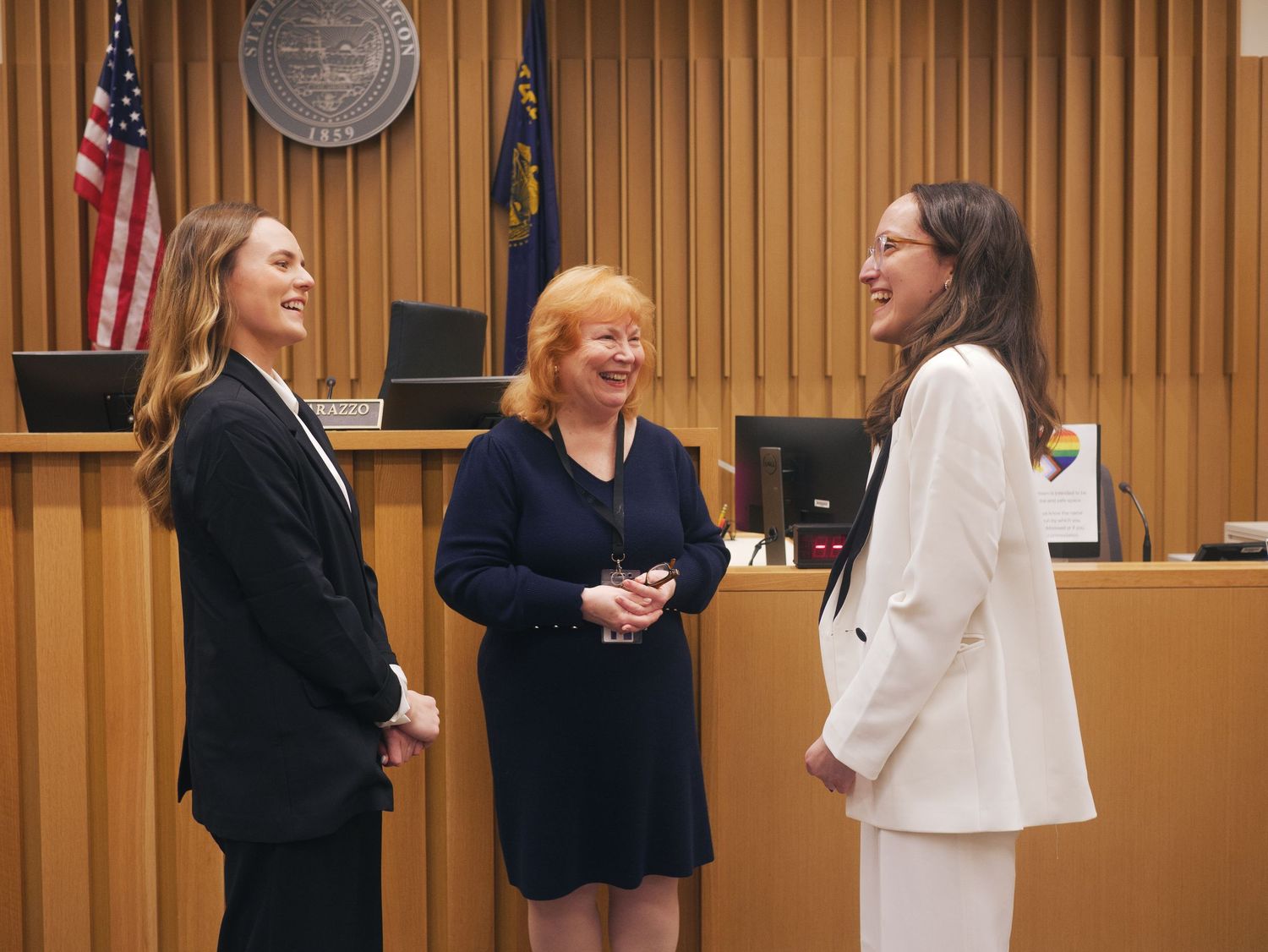 Three women share a joyful conversation inside a courtroom with an American flag backdrop.