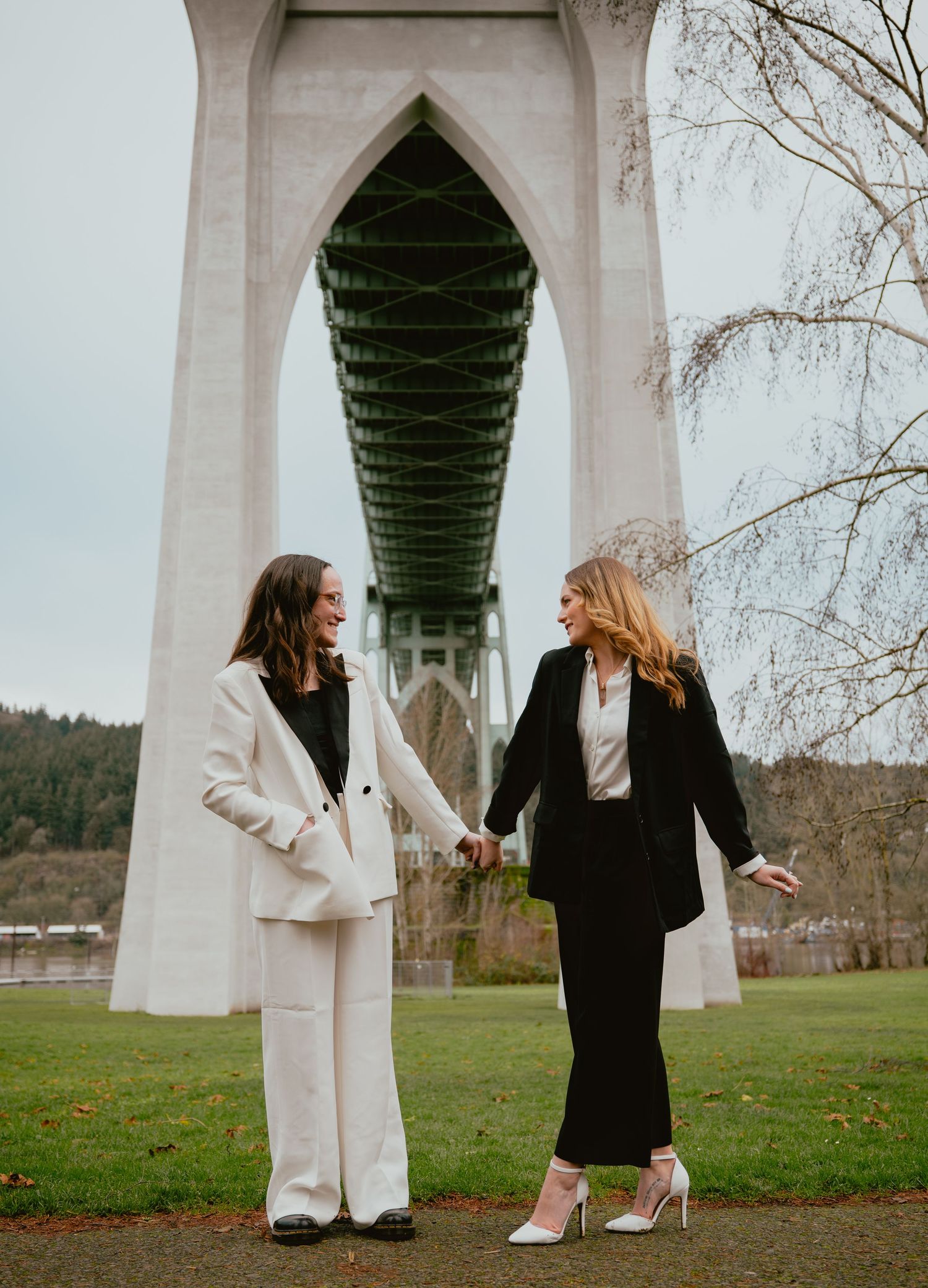 Two women in black and white suits holding hands and laughing under a large bridge.
