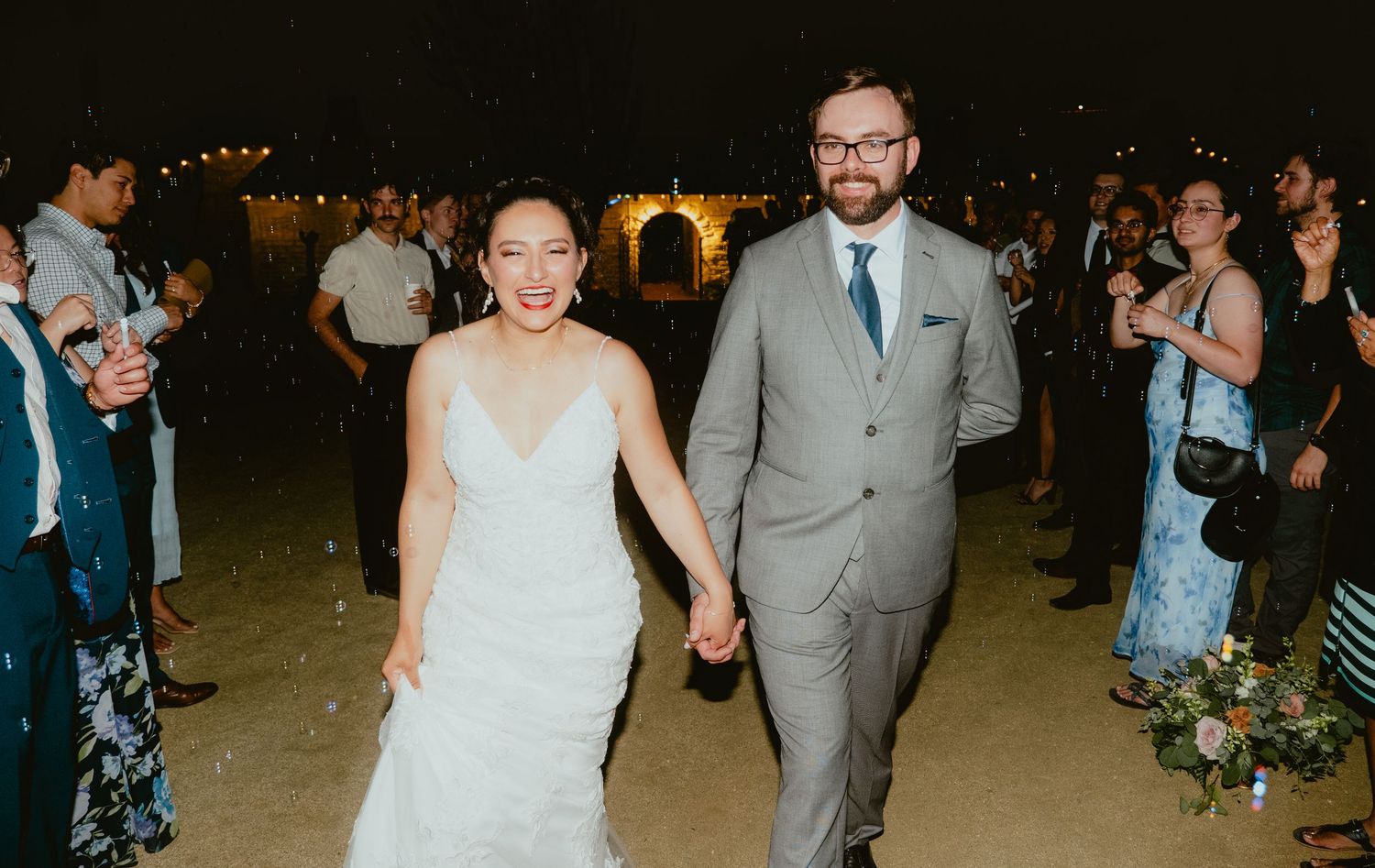 Smiling newlywed couple walking hand-in-hand at night with guests celebrating in the background.