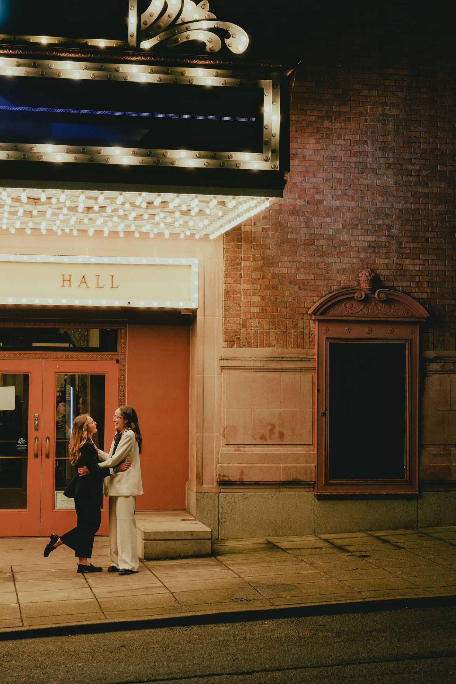 Two women share a romantic moment outside a concert hall entrance at night.