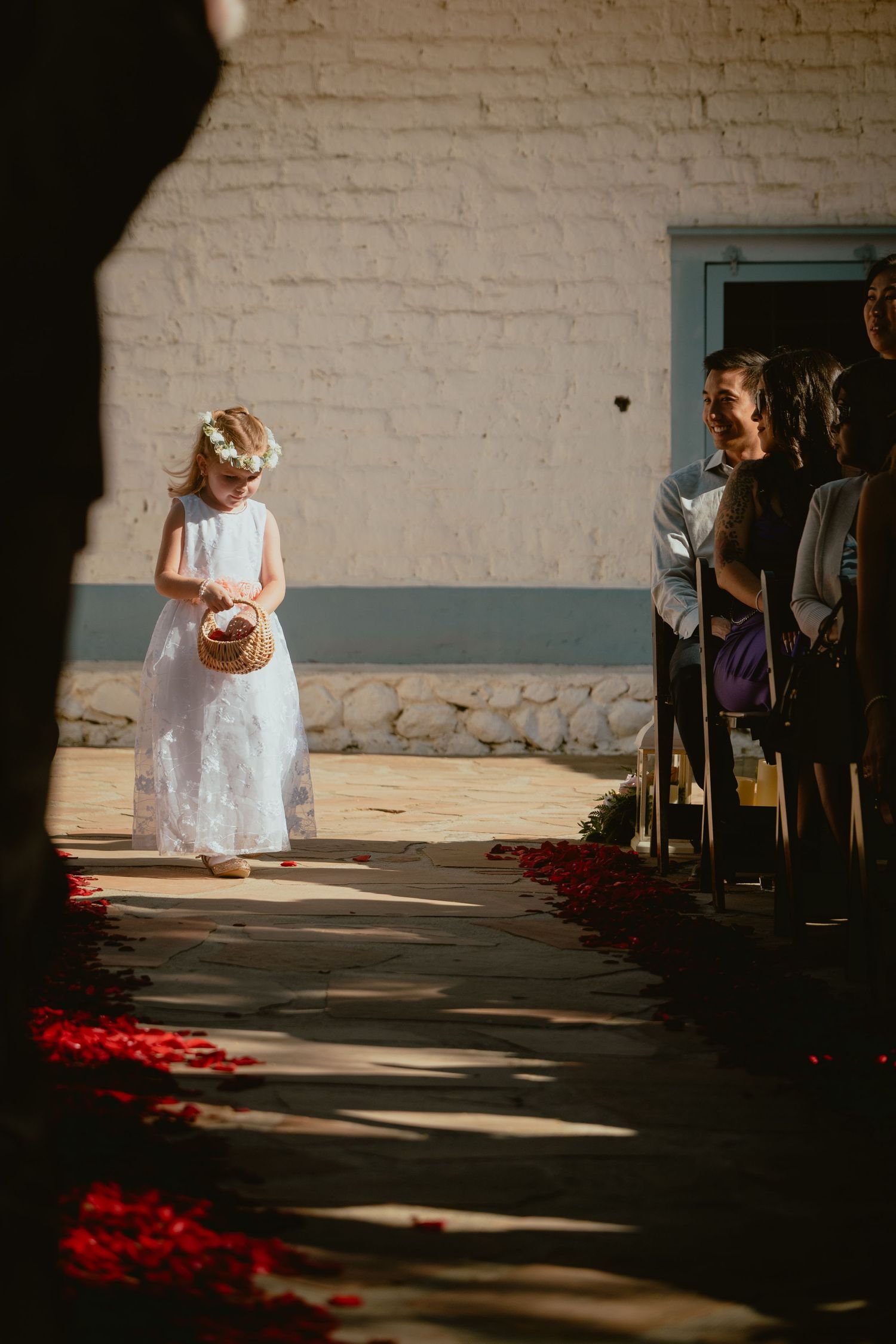 Flower girl in white dress walking down rose petal aisle at wedding.