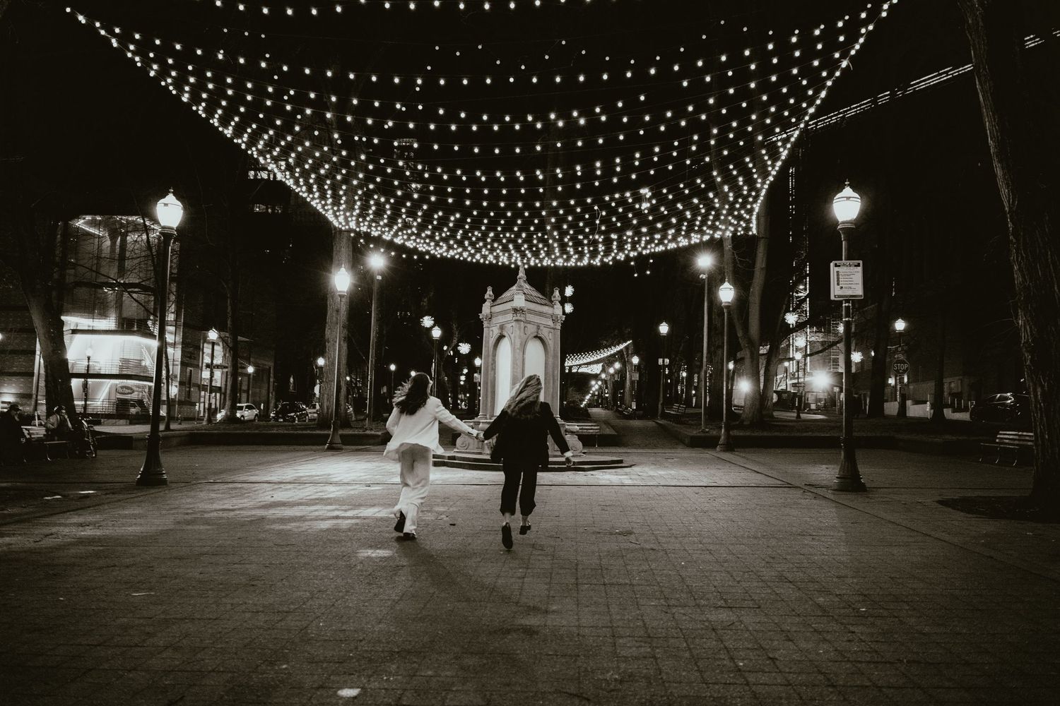 Black and white photo of couple running hand-in-hand under canopy of string lights in city square at night.